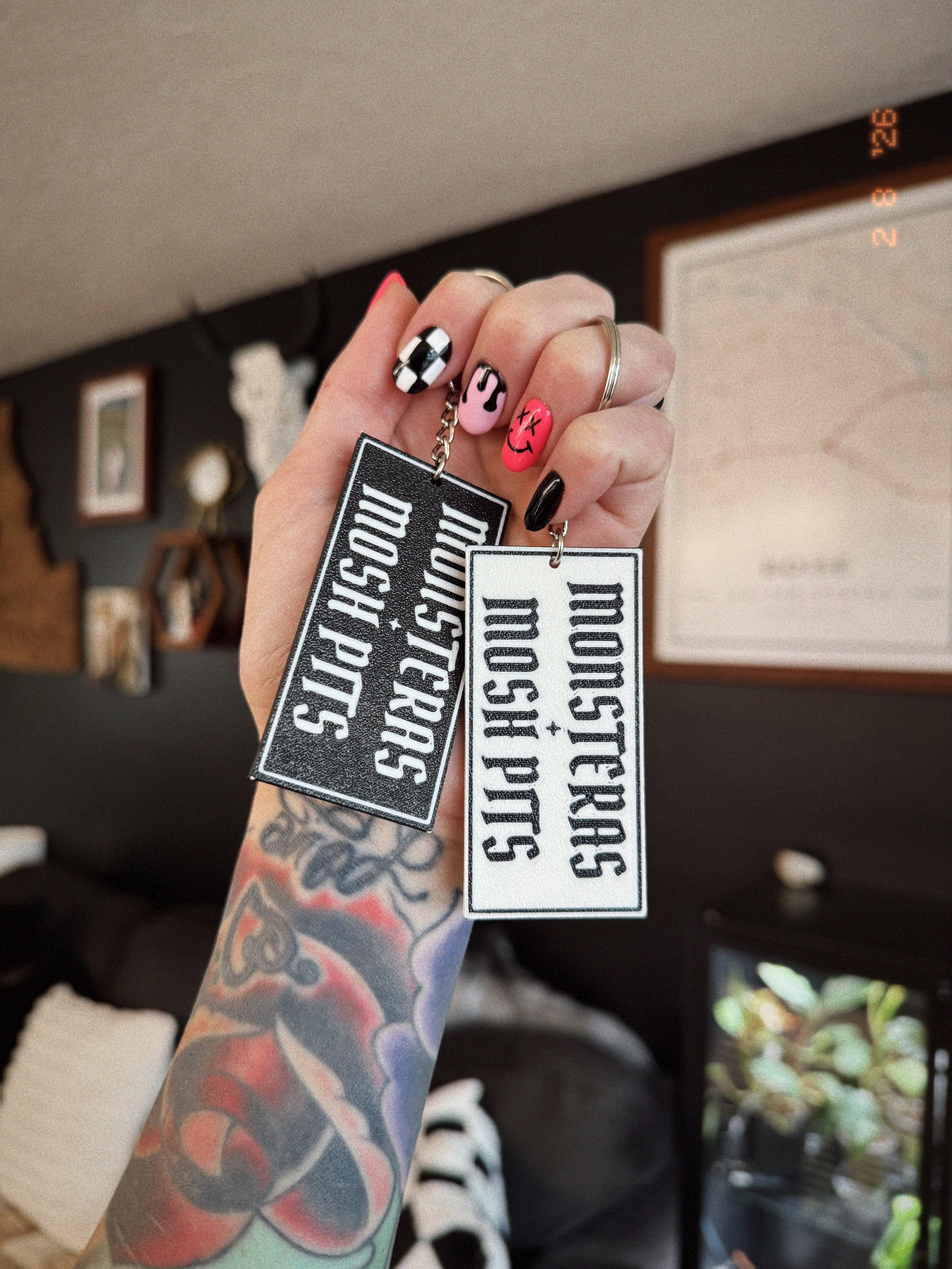 Hand holding two Monsteras + Mosh Pits 3D printed keychains in black and white with keychain clips.