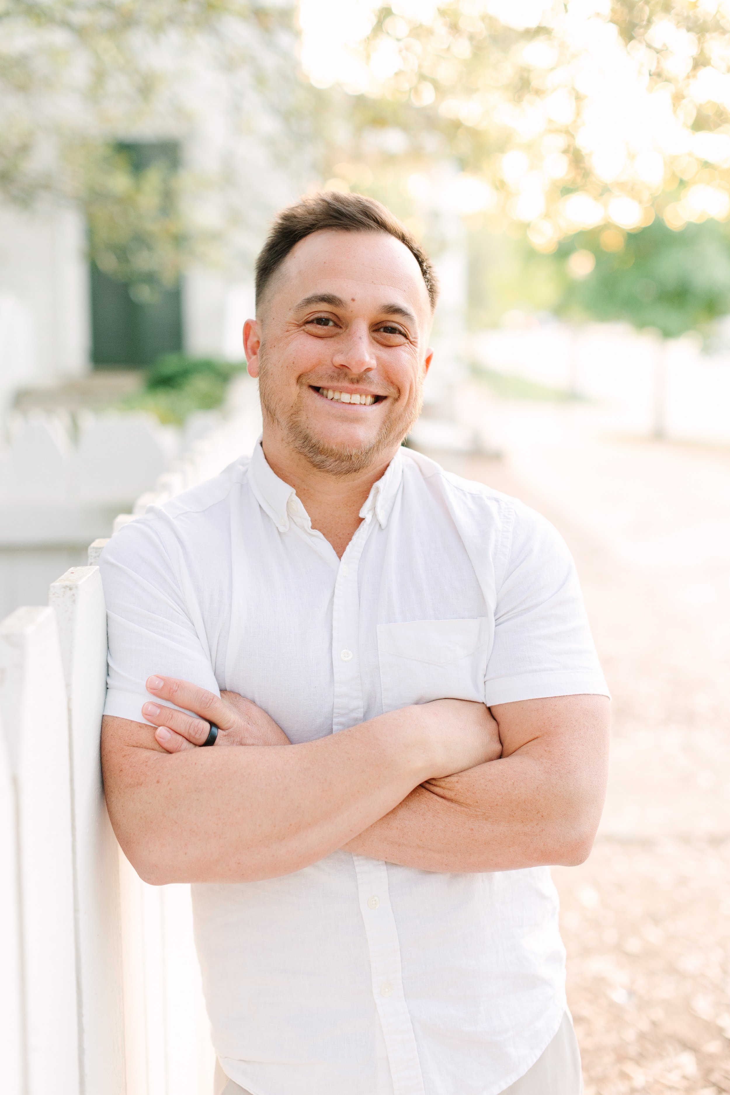 Smiling man with crossed arms wearing a white shirt, standing outdoors near a white fence, with greenery and trees in the background during daylight.