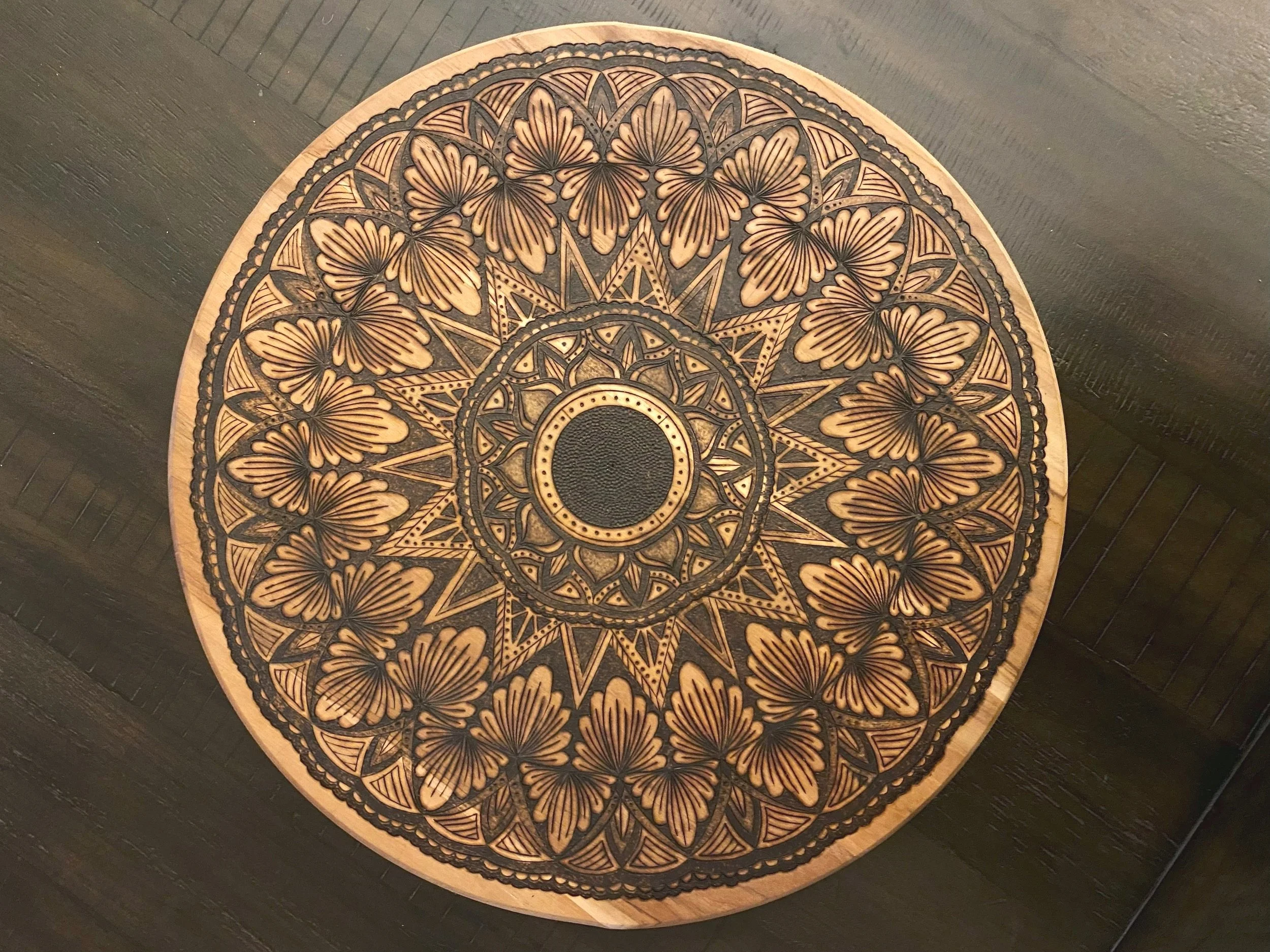 A round decorative wooden tray with intricate black designs featuring butterflies and geometric patterns, placed on a dark wooden surface.