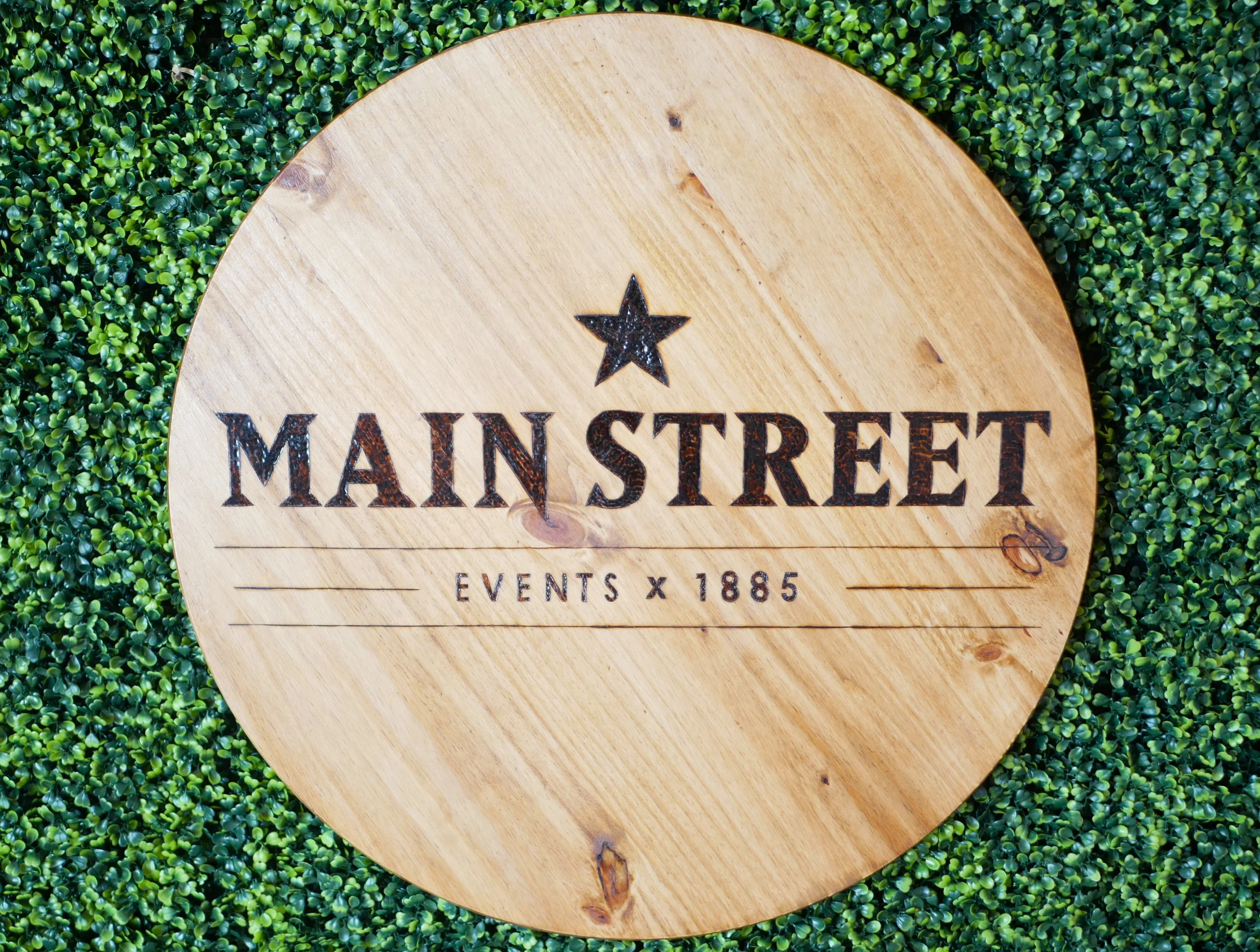 A round wooden sign engraved with 'Main Street Events x 1885' and a star, surrounded by green artificial grass.
