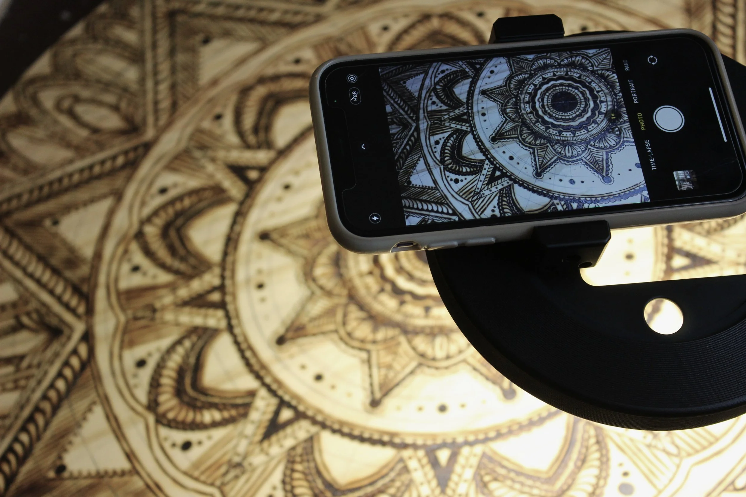 A smartphone on a tripod takes a picture of an intricate mandala design on a wooden surface.