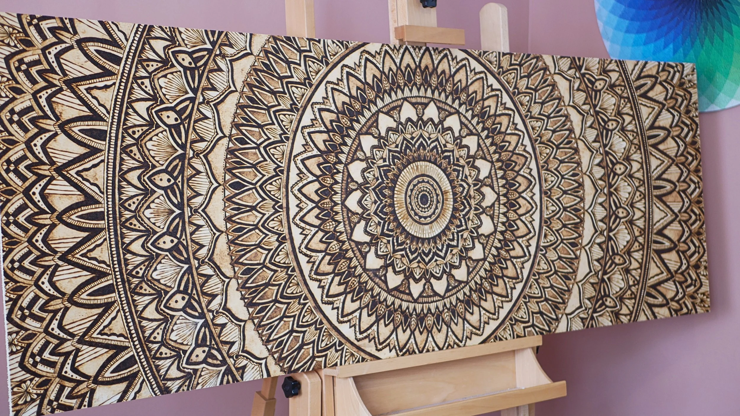 A large canvas with an intricate, symmetrical black and tan mandala design is mounted on a wooden easel against a pink wall. To the right, part of a colorful paper lantern is visible.