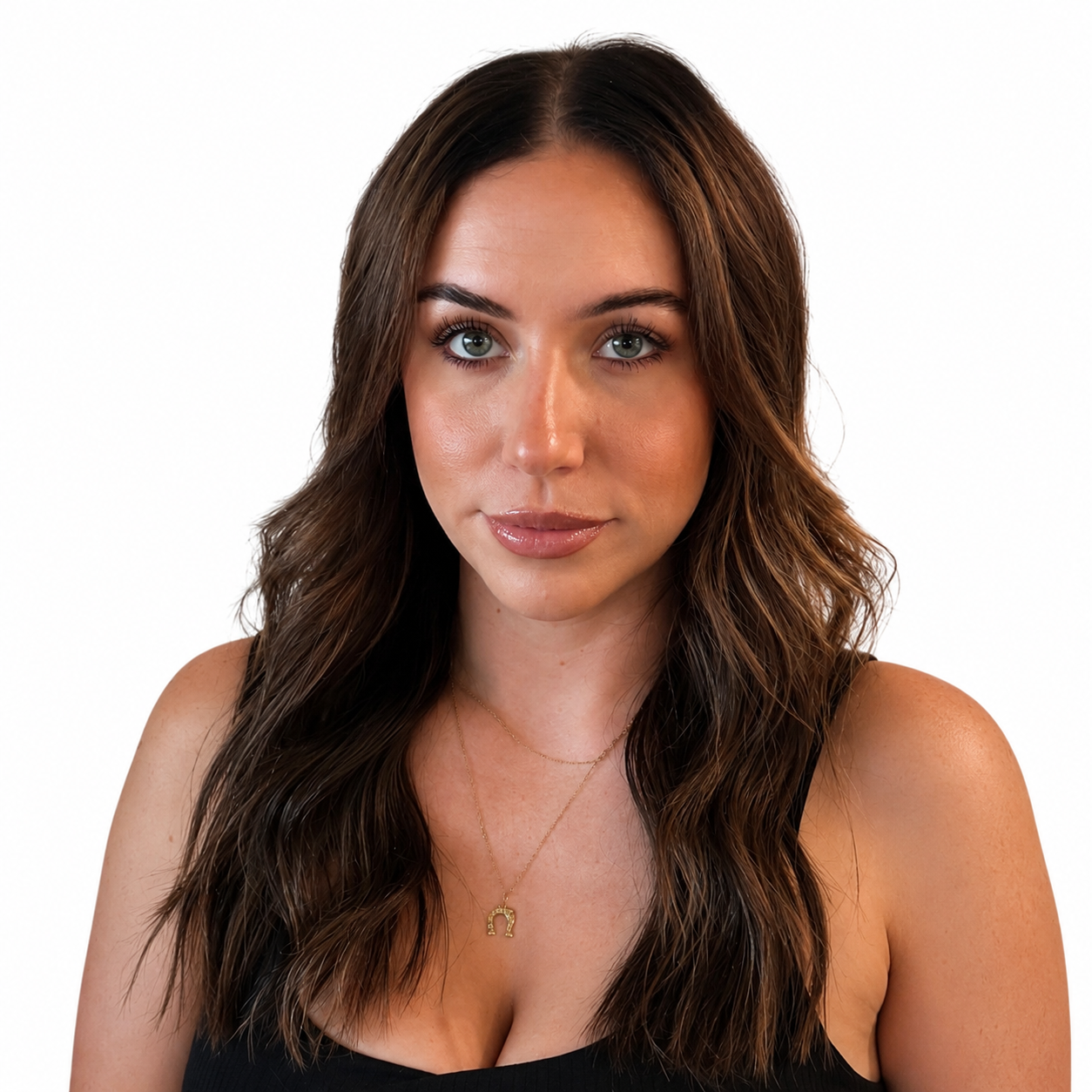 Close-up portrait of a woman with long wavy brown hair, light makeup, blue eyes, wearing a black top and a gold necklace with a horseshoe pendant, posing against a plain white background.