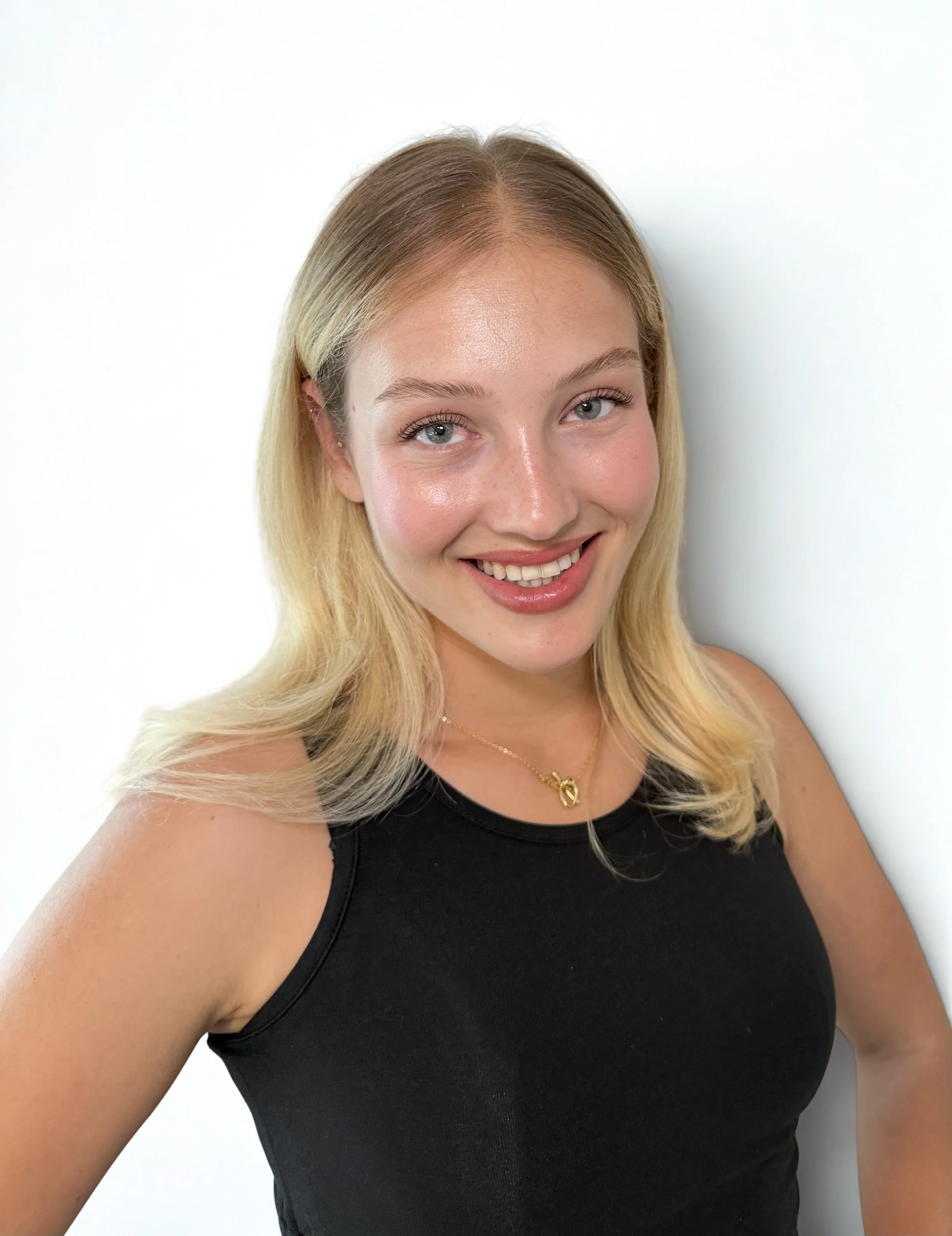 A young woman with long blonde hair, light skin, and blue eyes, smiling at the camera, wearing a black sleeveless top and a gold necklace with a heart-shaped pendant, standing against a plain white background.
