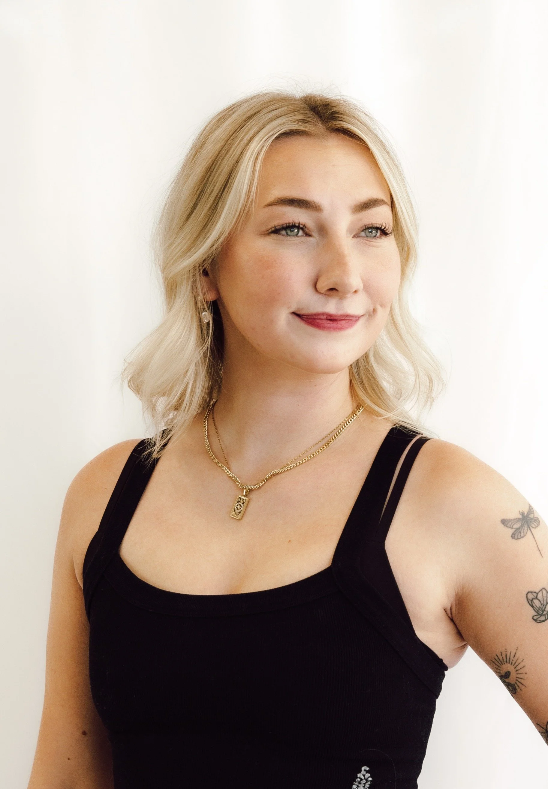 Portrait of a young woman with blonde hair, wearing a black sleeveless top, several necklaces, and earrings, standing against a plain white background.