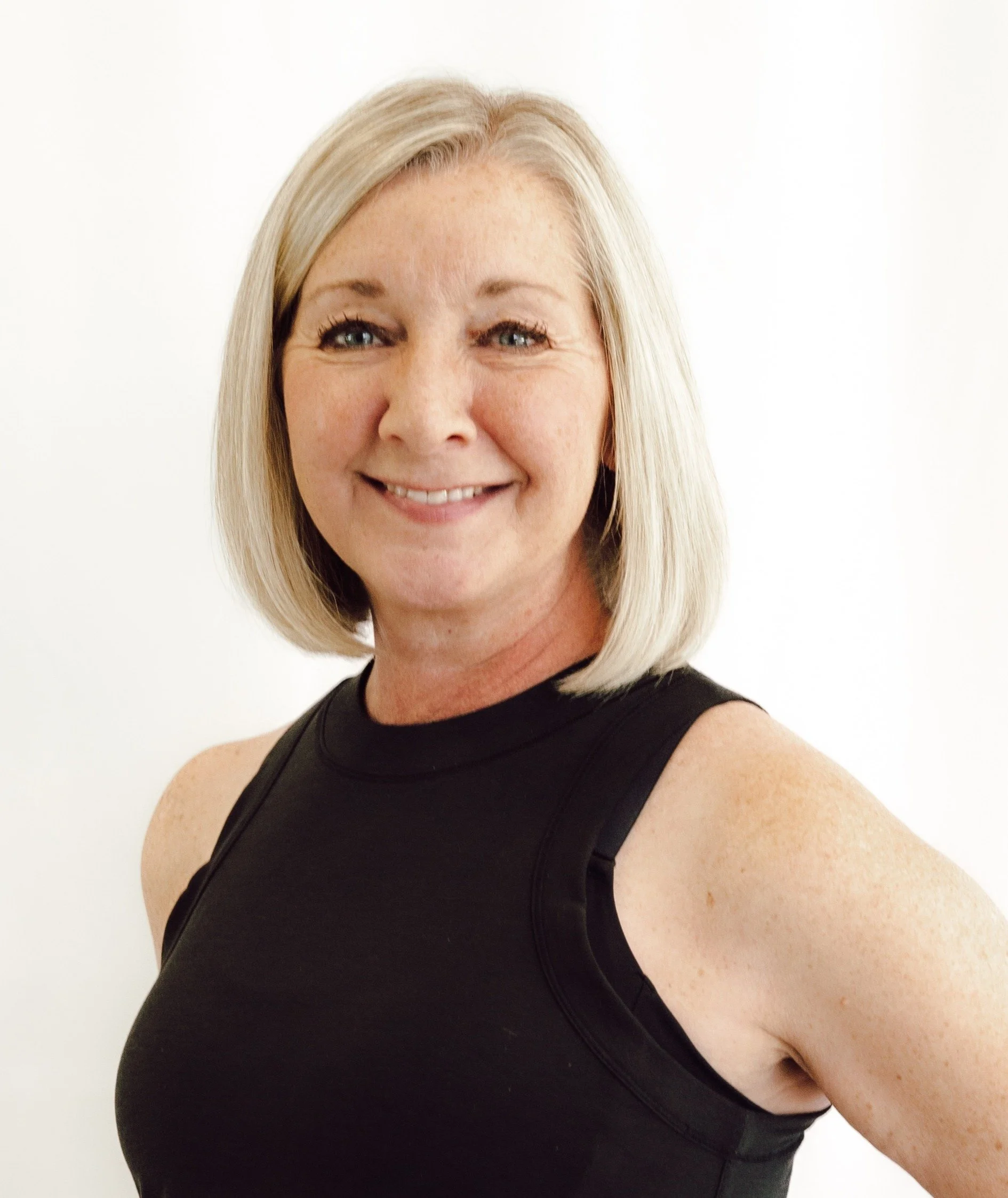Close-up of a woman with blonde hair, blue eyes, and freckles, smiling while wearing a black sleeveless top, against a plain white background.