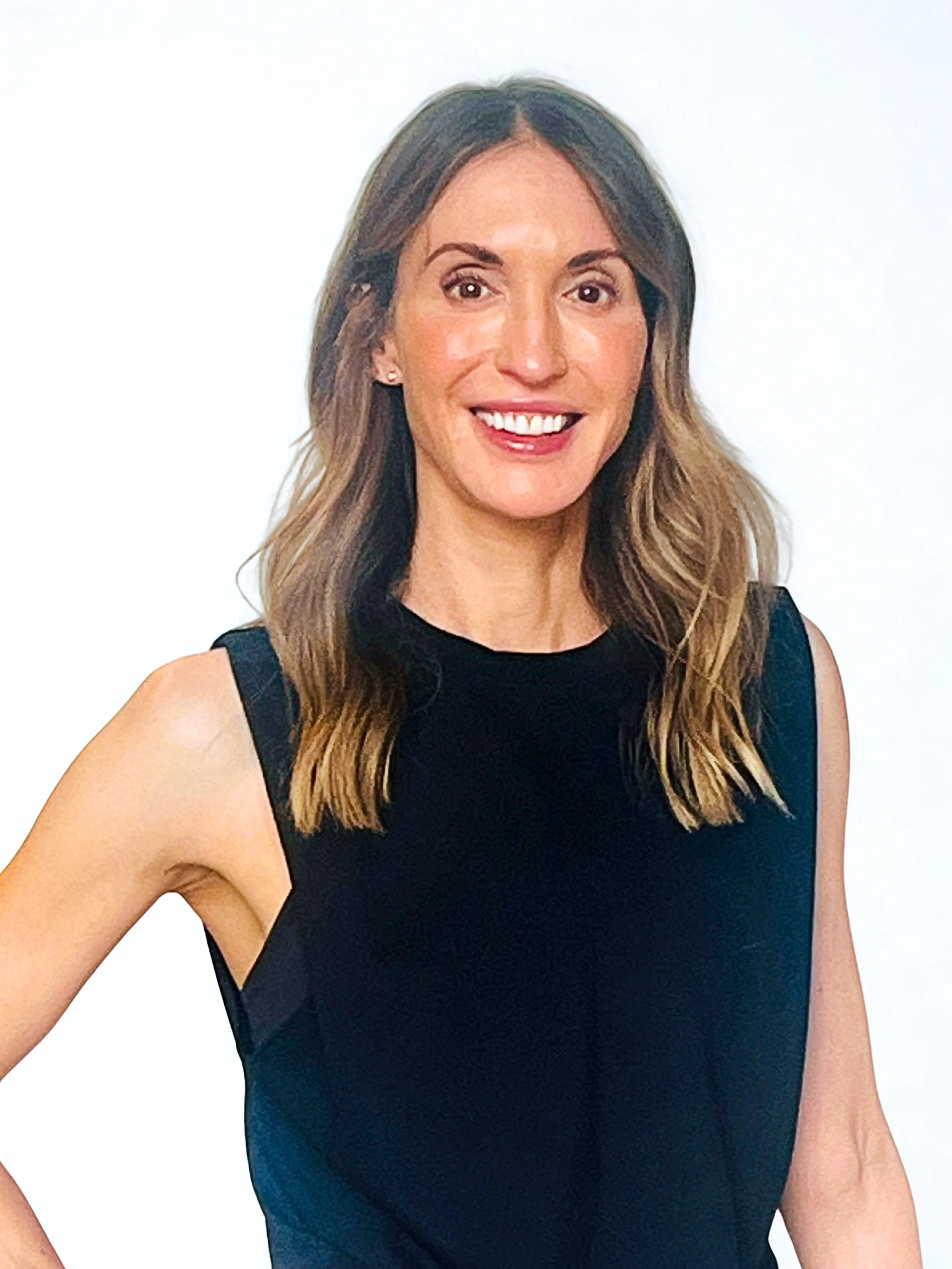 A woman with long, wavy brown hair, wearing a sleeveless black top, smiling with white teeth, standing against a plain white background.