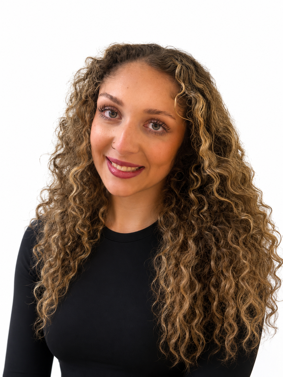 A young woman with curly hair, wearing a black top, smiling and looking at the camera against a white background.
