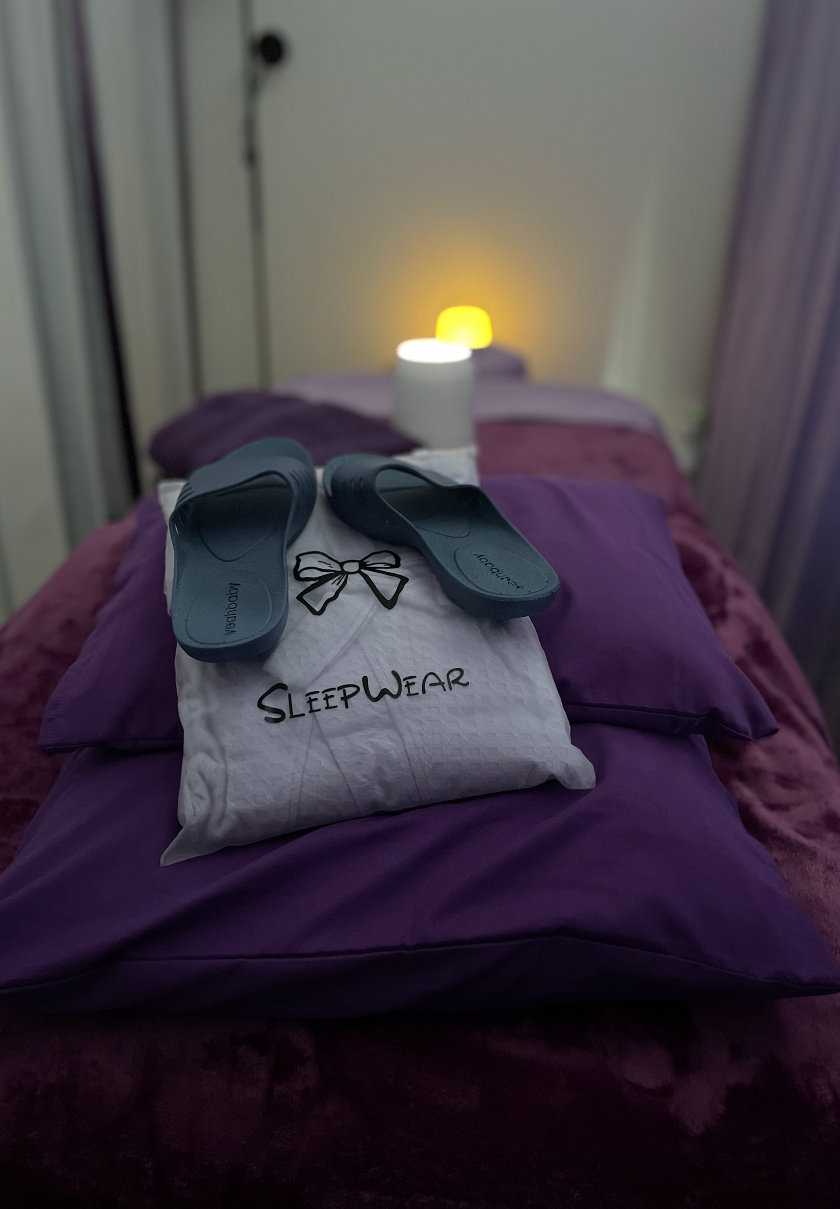 A bed with purple sheets. On the bed, there is a white sleepwear package with black text, a pair of blue slides, and a cloth with a bow design. In the background, there are two lamps emitting yellow and white light, and an adjustable wall lamp is att