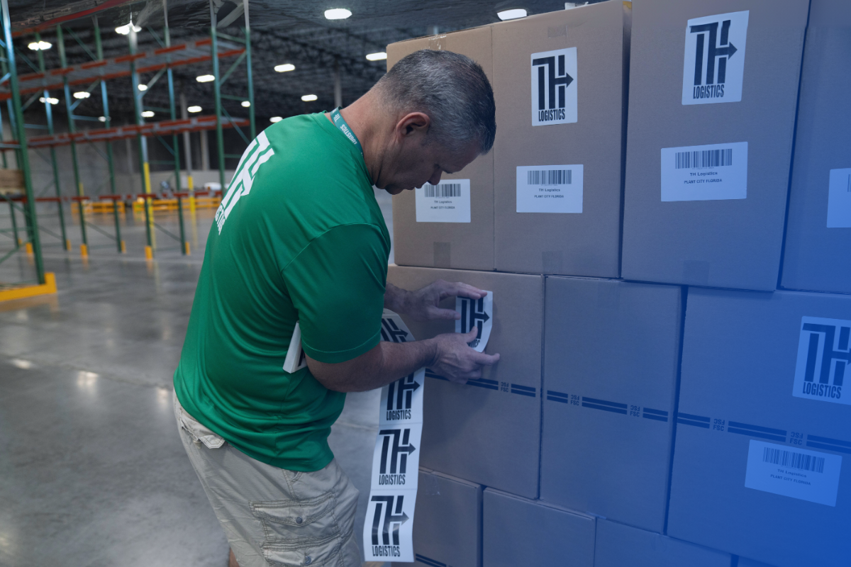 Worker in a green TH Logistics shirt applying branded labels to stacked cardboard boxes in a warehouse. Organized packing operations, attention to detail, and professional fulfillment processes within a spacious distribution center.
