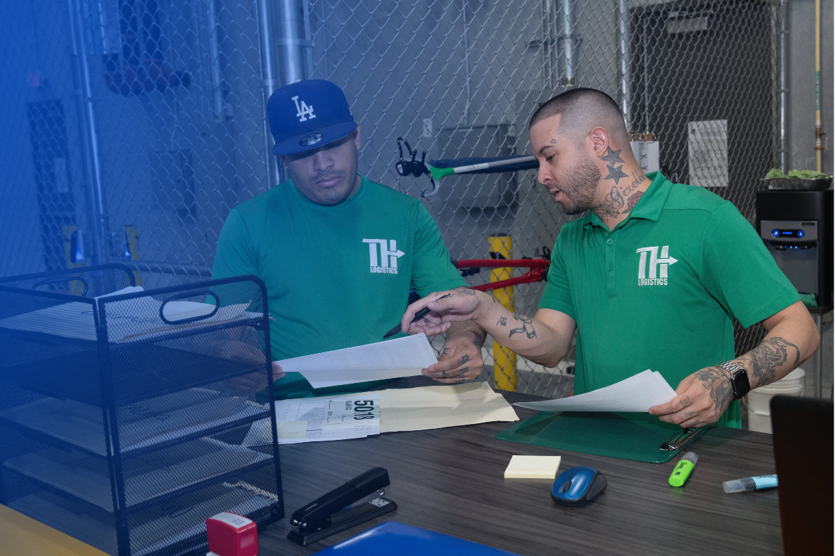 Two TH Logistics team members in green uniforms reviewing documents at a work station inside a secure facility. . Surrounded by packing supplies and office tools,  they collaborate on shipment or inventory paperwork.