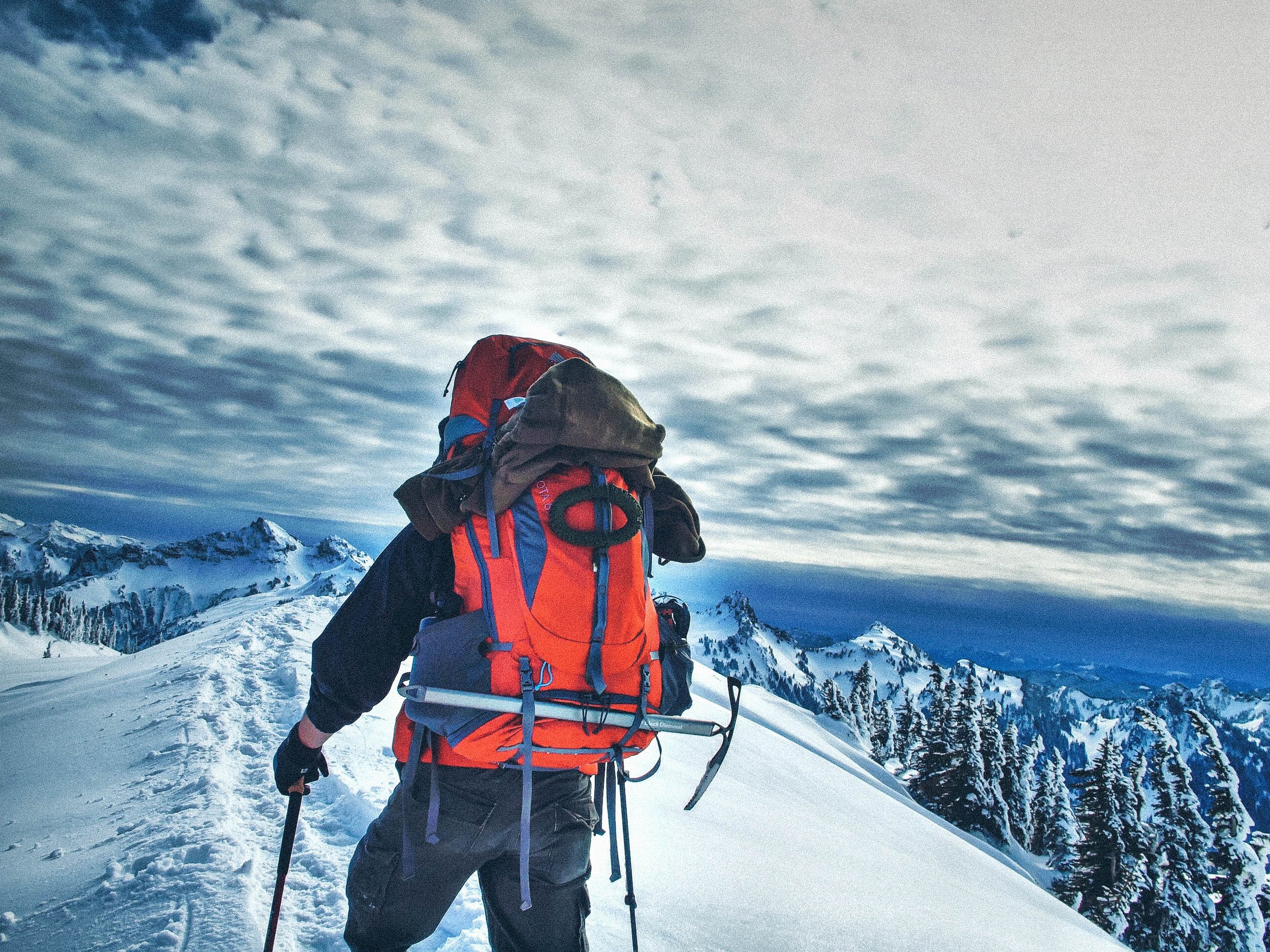 A person dressed in winter gear, including a red and black jacket and backpack, is snowshoeing on a snowy mountain trail with snow-covered peaks in the background.