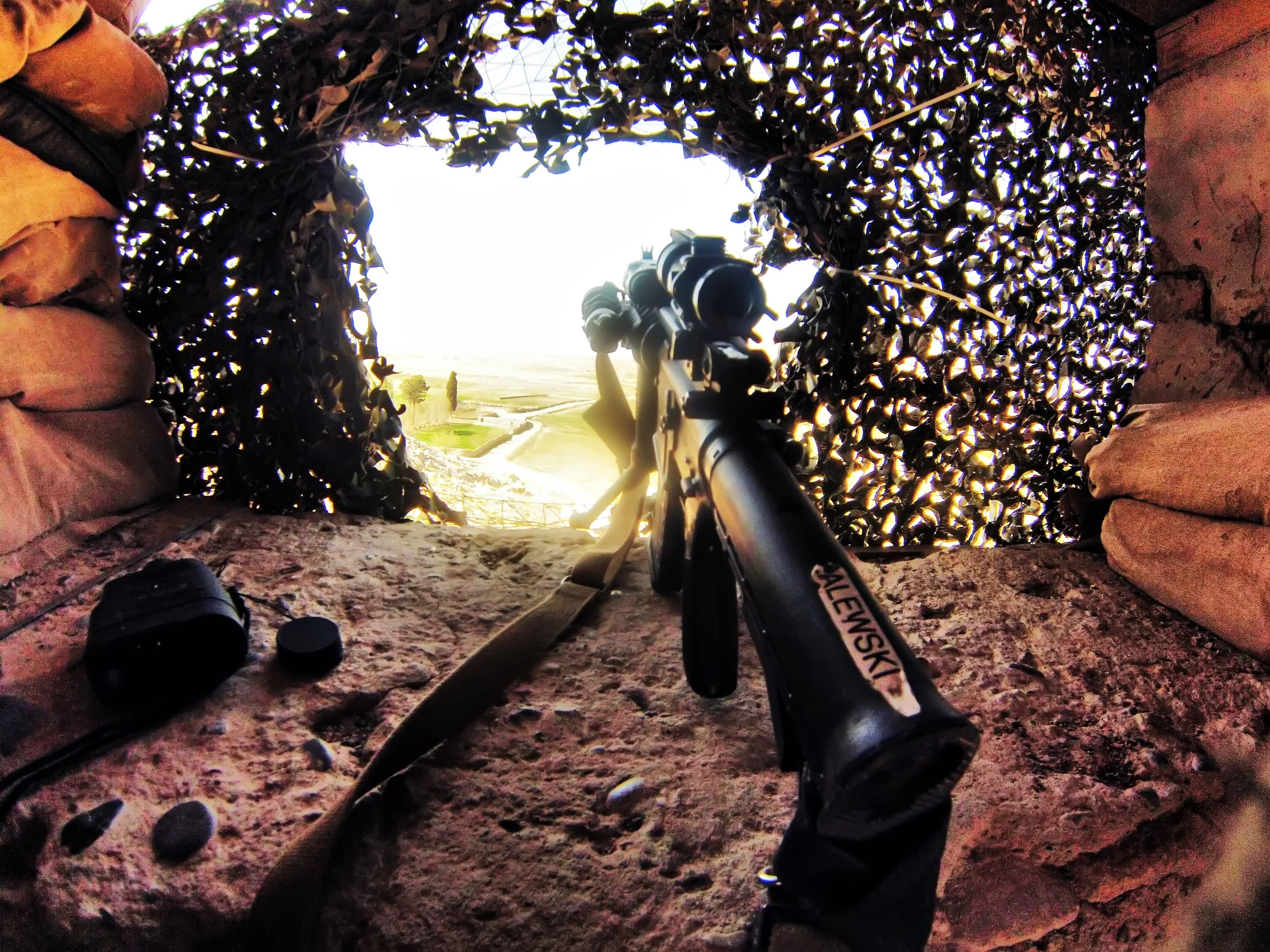 View through a small, stone-walled observation opening with camouflage netting, featuring a scoped rifle pointed outward, overlooking a landscape with fields and trees.