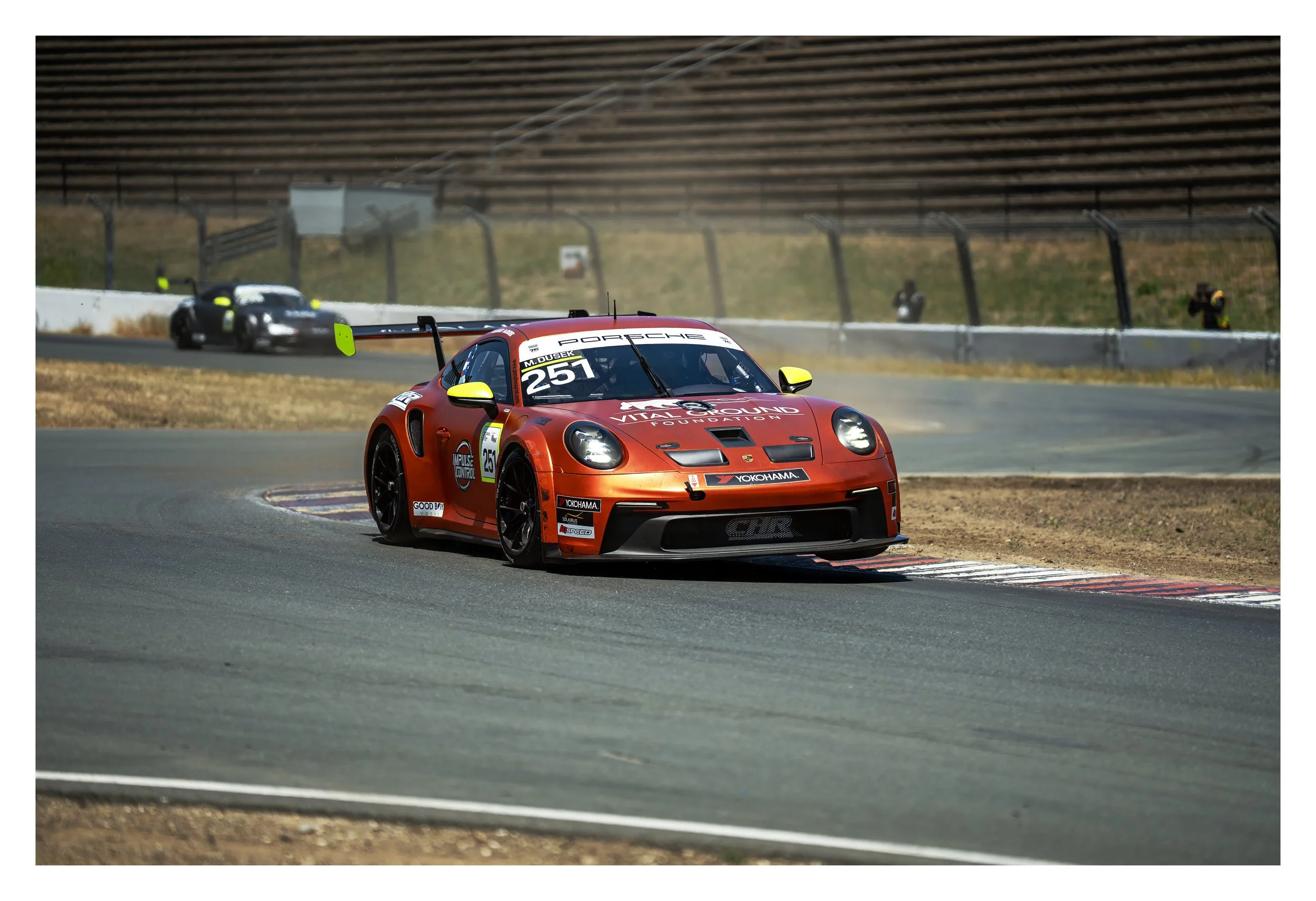 Red Porsche race car on a racetrack with a black sports car in the background and a driver on the right side of the track with a camera.