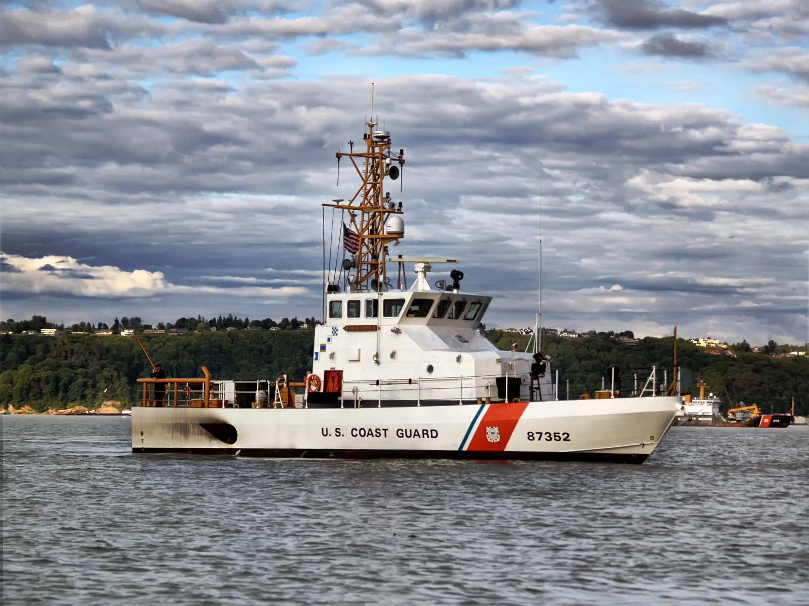 U.S. Coast Guard patrol boat on the water with a cloudy sky and hilly landscape in the background.