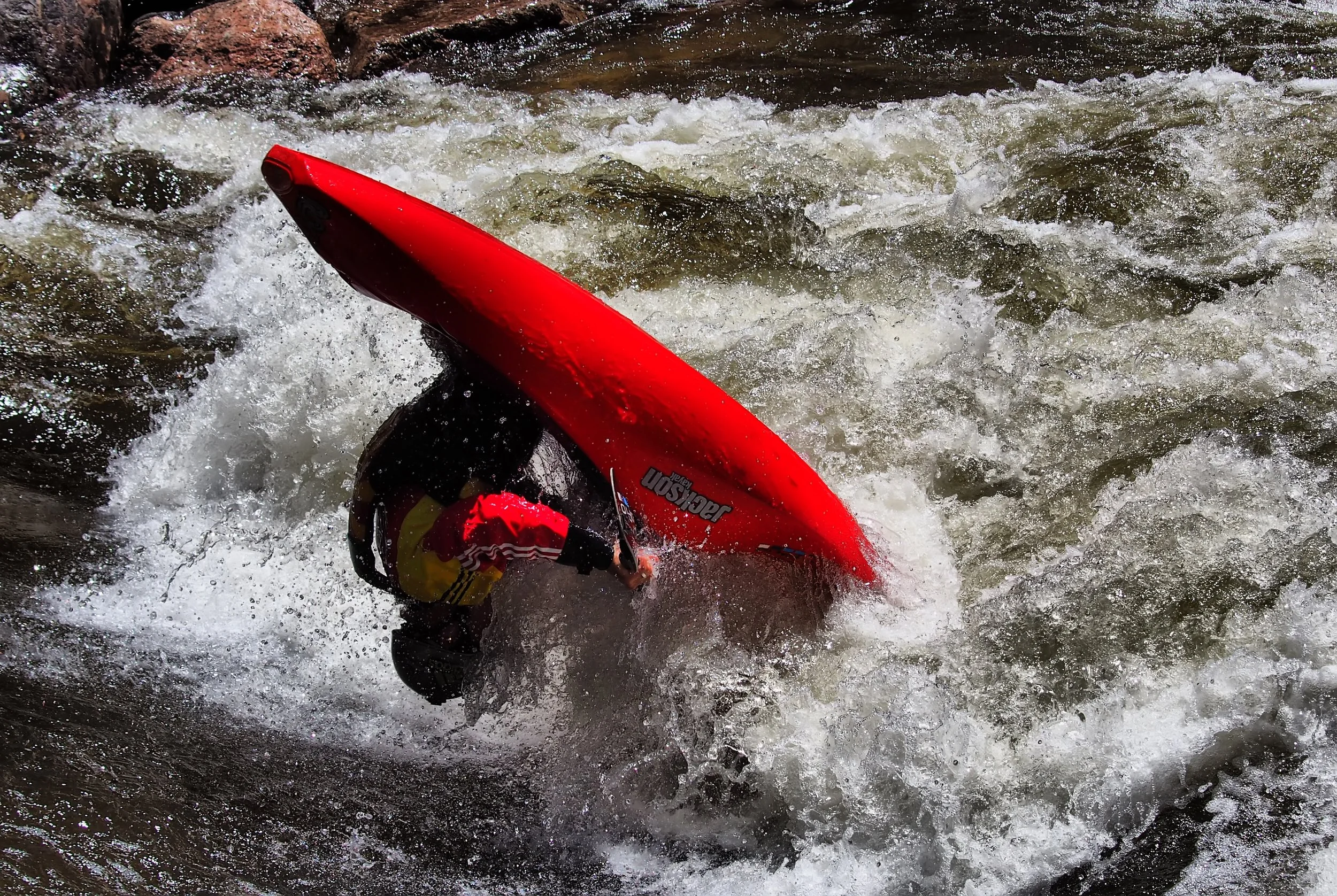 A person in a yellow and black life jacket kayaking through white water rapids in a river, with a red kayak and partially submerged. The person's head is bent down, and the scene is full of splashing water and rocks.