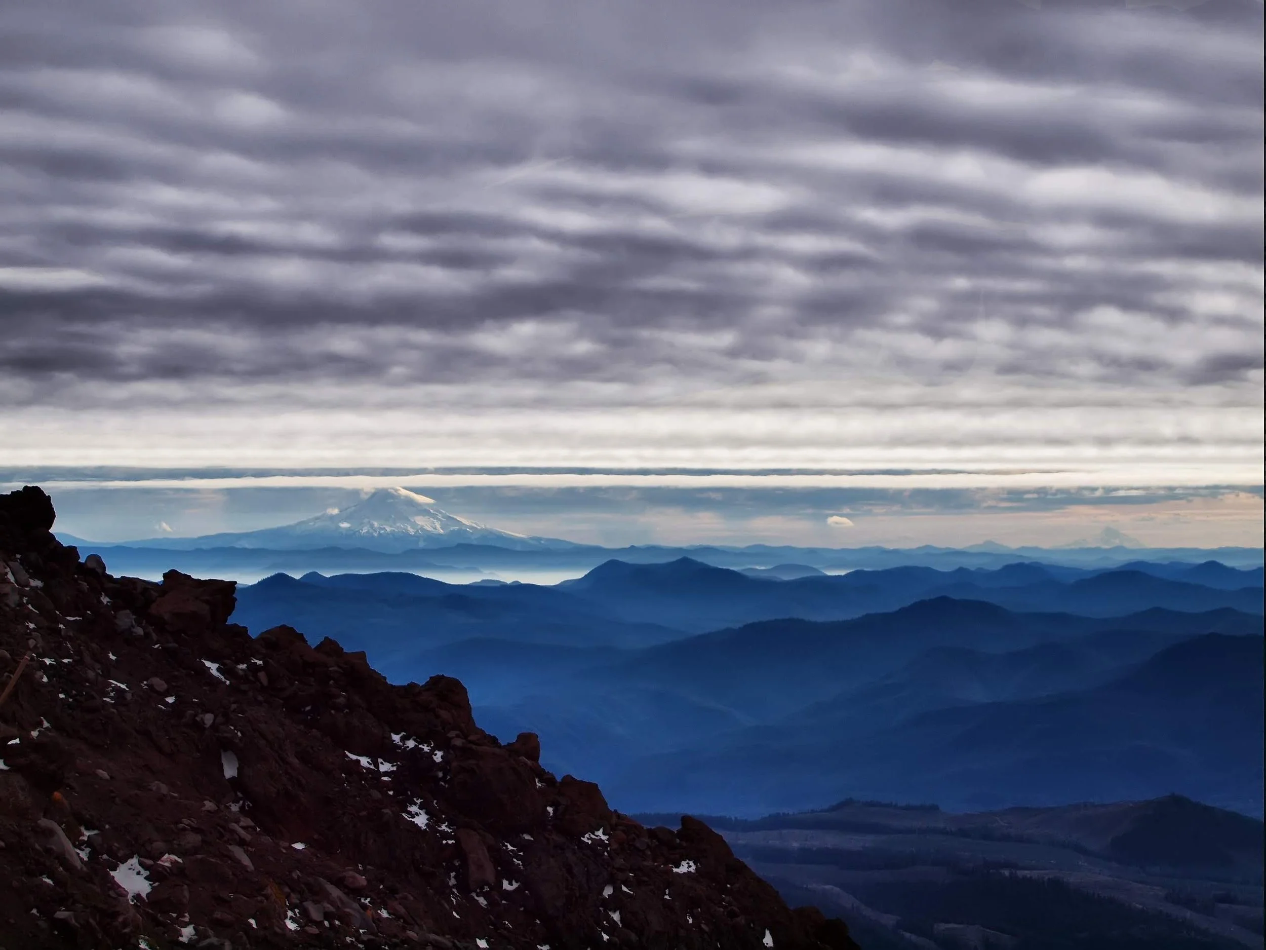A mountain landscape with dark, rocky slopes in the foreground, layers of mountain ridges in the middle ground, and a snow-capped volcano in the distance under a cloudy sky.