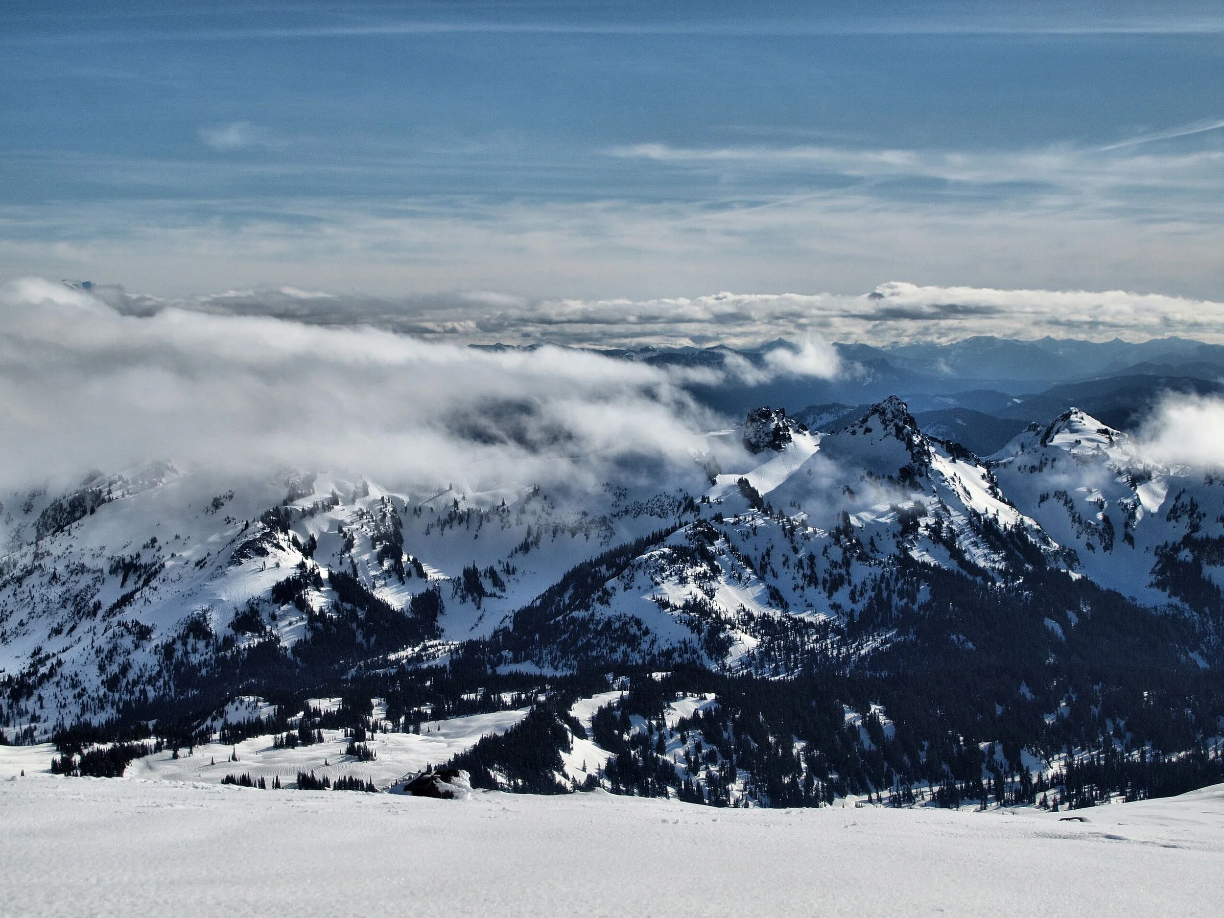 Snow-covered mountain range with rugged peaks and scattered trees, under a partly cloudy sky with layers of clouds and blue sky above.