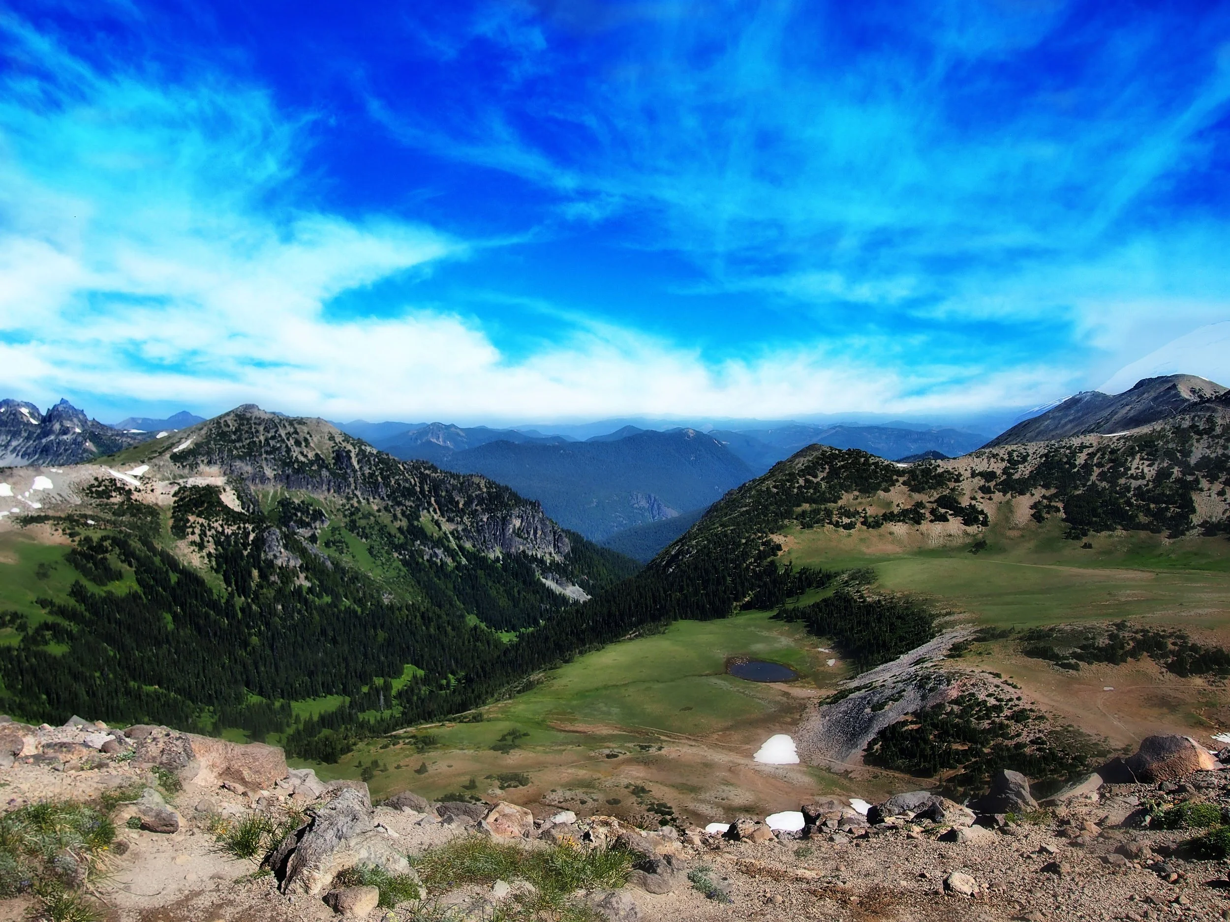 Scenic mountain landscape with green valleys, rocky slopes, scattered patches of snow, and a blue sky with wispy clouds.