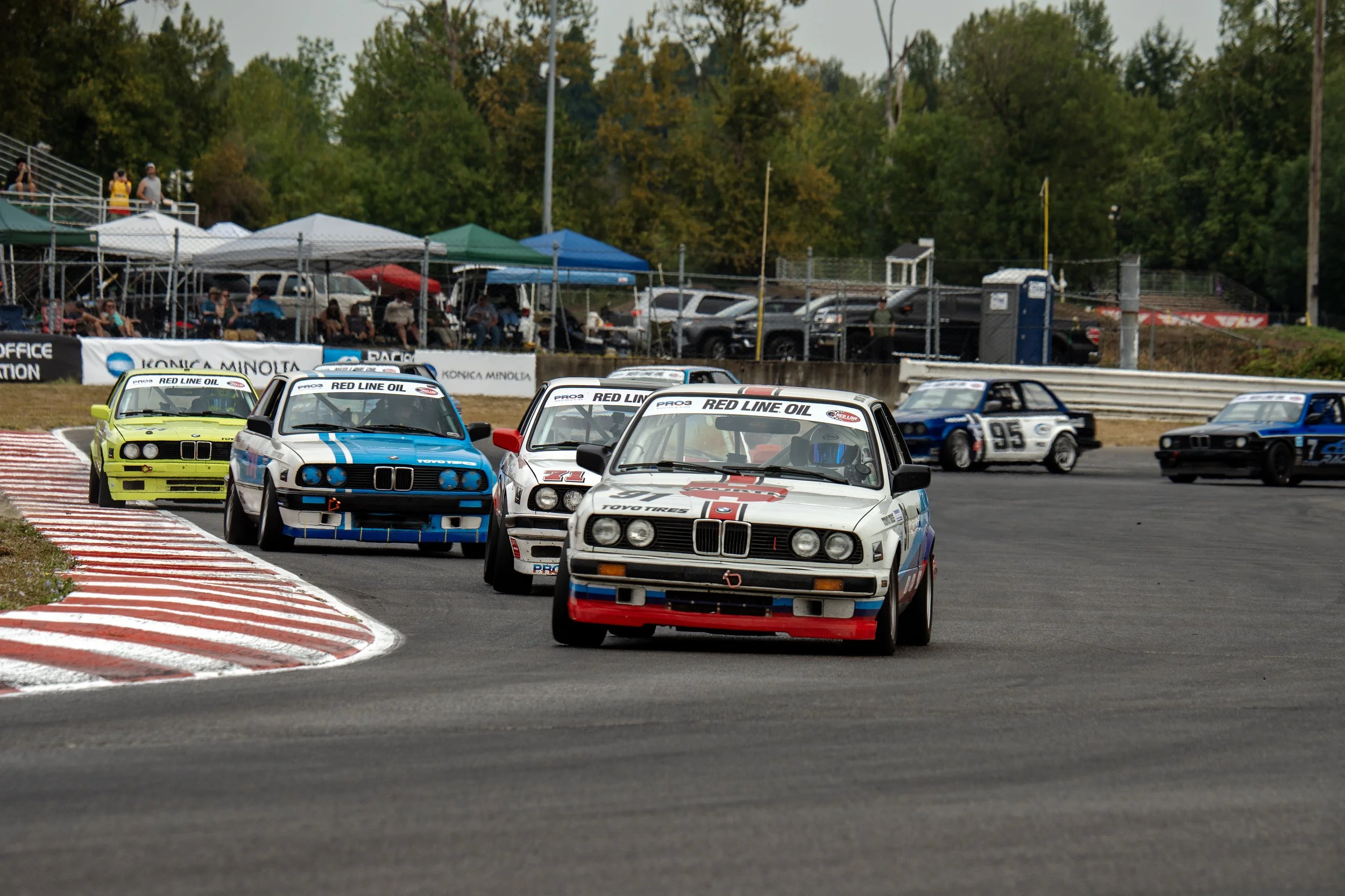 Several race cars on a track during a motorsport event, with spectators and parked cars in the background under a partly cloudy sky.
