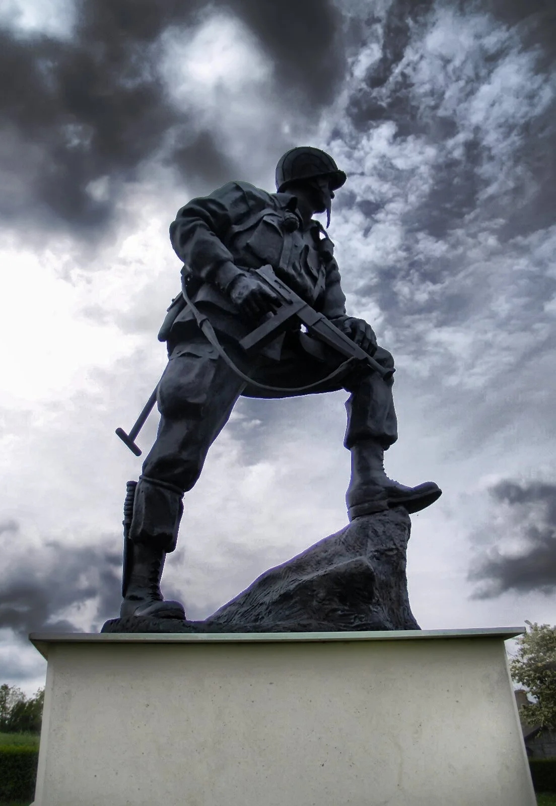 Soldier statue in uniform holding a rifle, standing with one foot on a rock, against a cloudy sky.