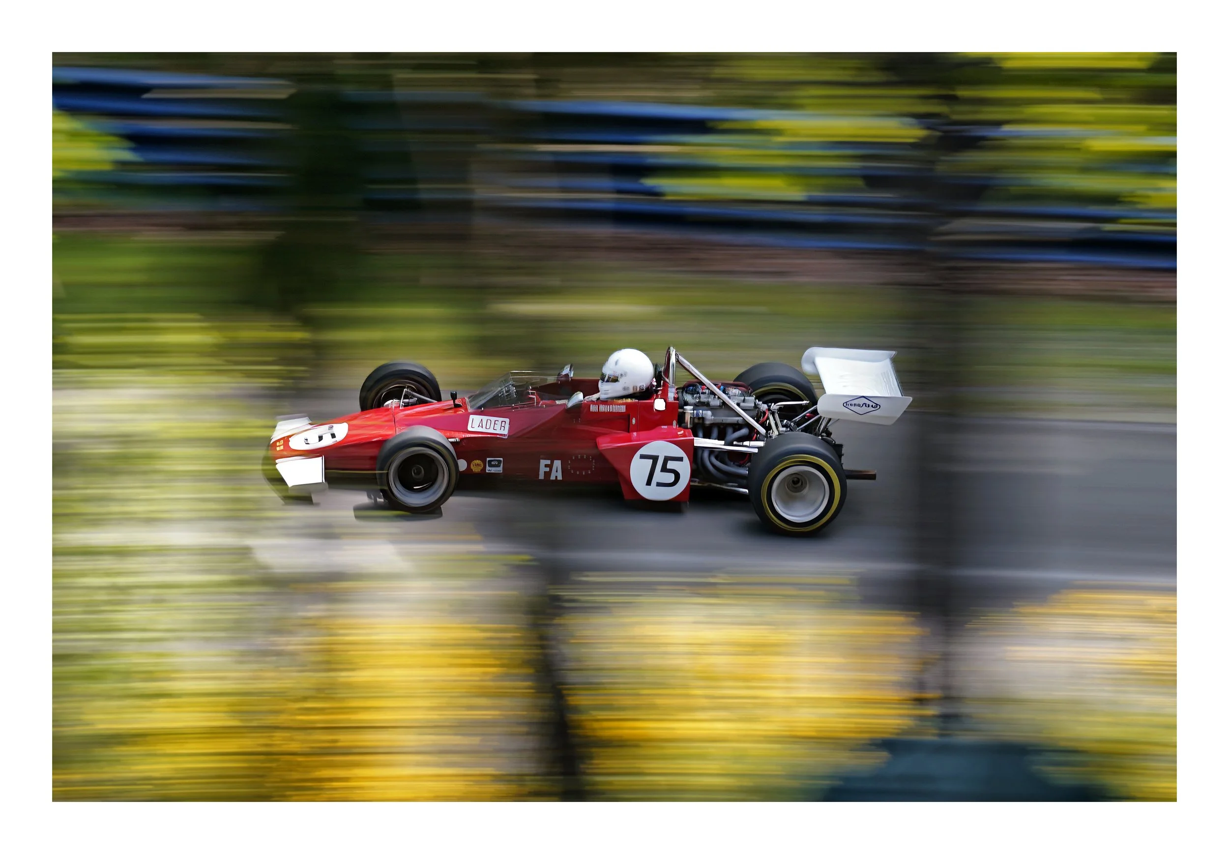 A red race car with the number 75 is moving at high speed, with a blurred background of trees and greenery.