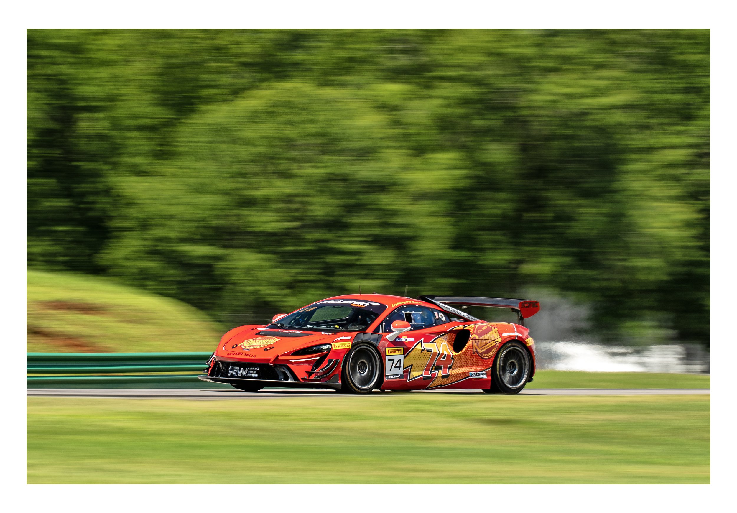Red race car with number 74 and various sponsor logos, speeding on a race track with green trees blurred in the background.