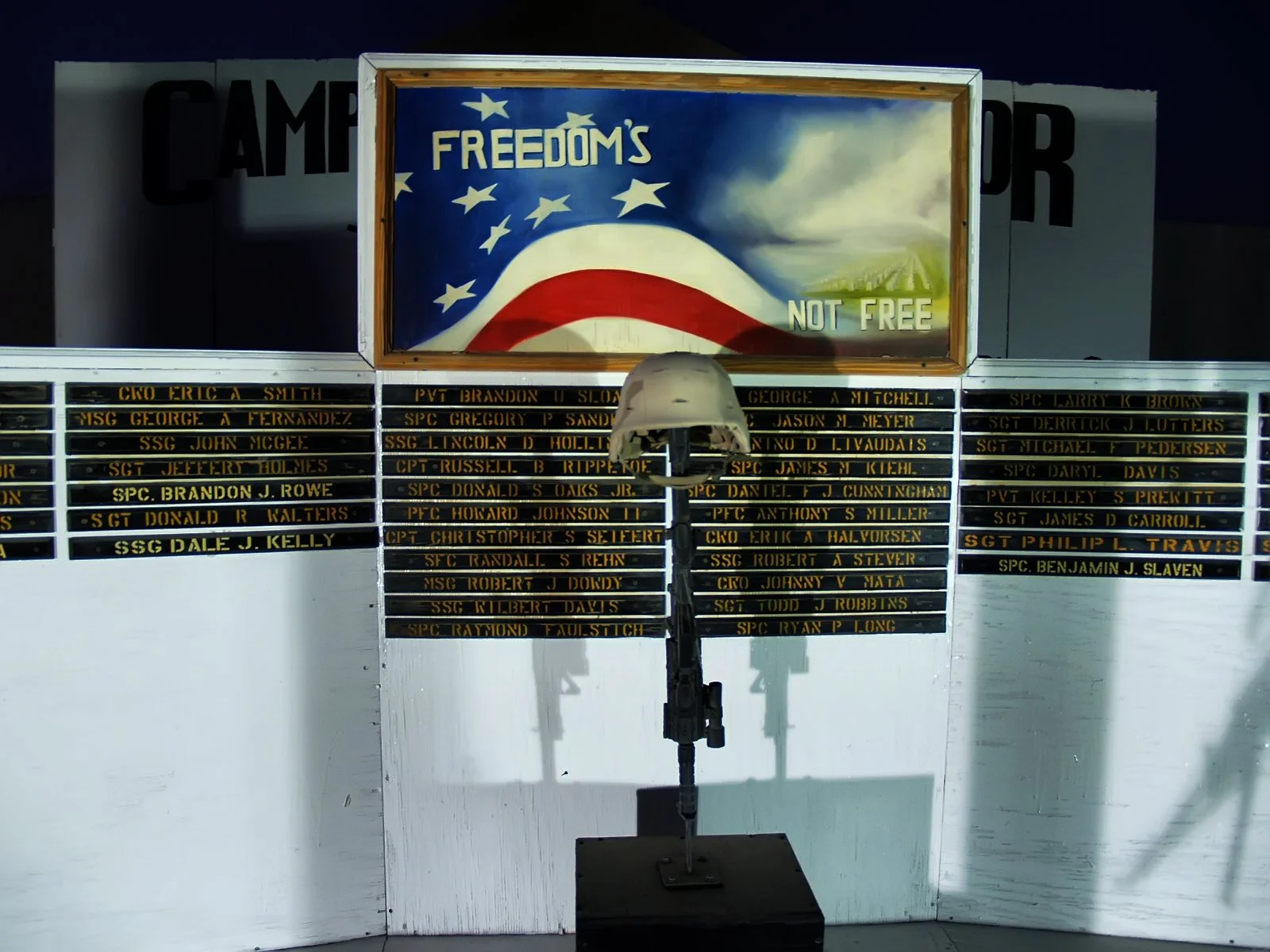 Memorial display with a helmet mounted on a stand. Behind it, black nameplates with gold lettering list names of fallen soldiers. Above, a painted banner features the American flag with the words 'FREEDOM'S NOT FREE' and clouds in the background.