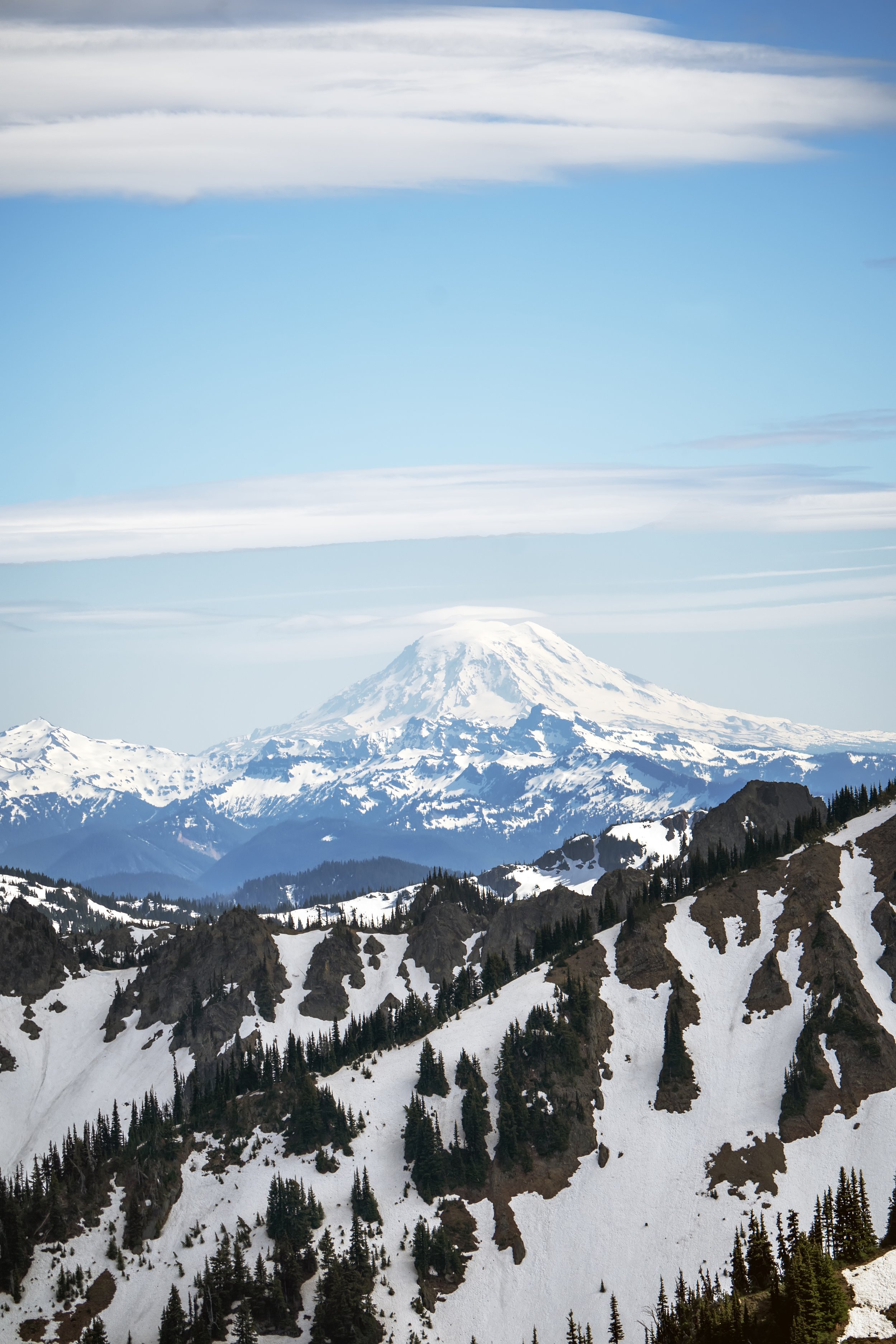 Snow-capped mountain, likely Mount Rainier, in a range with evergreen trees and rocky outcroppings in the foreground under a partly cloudy sky.