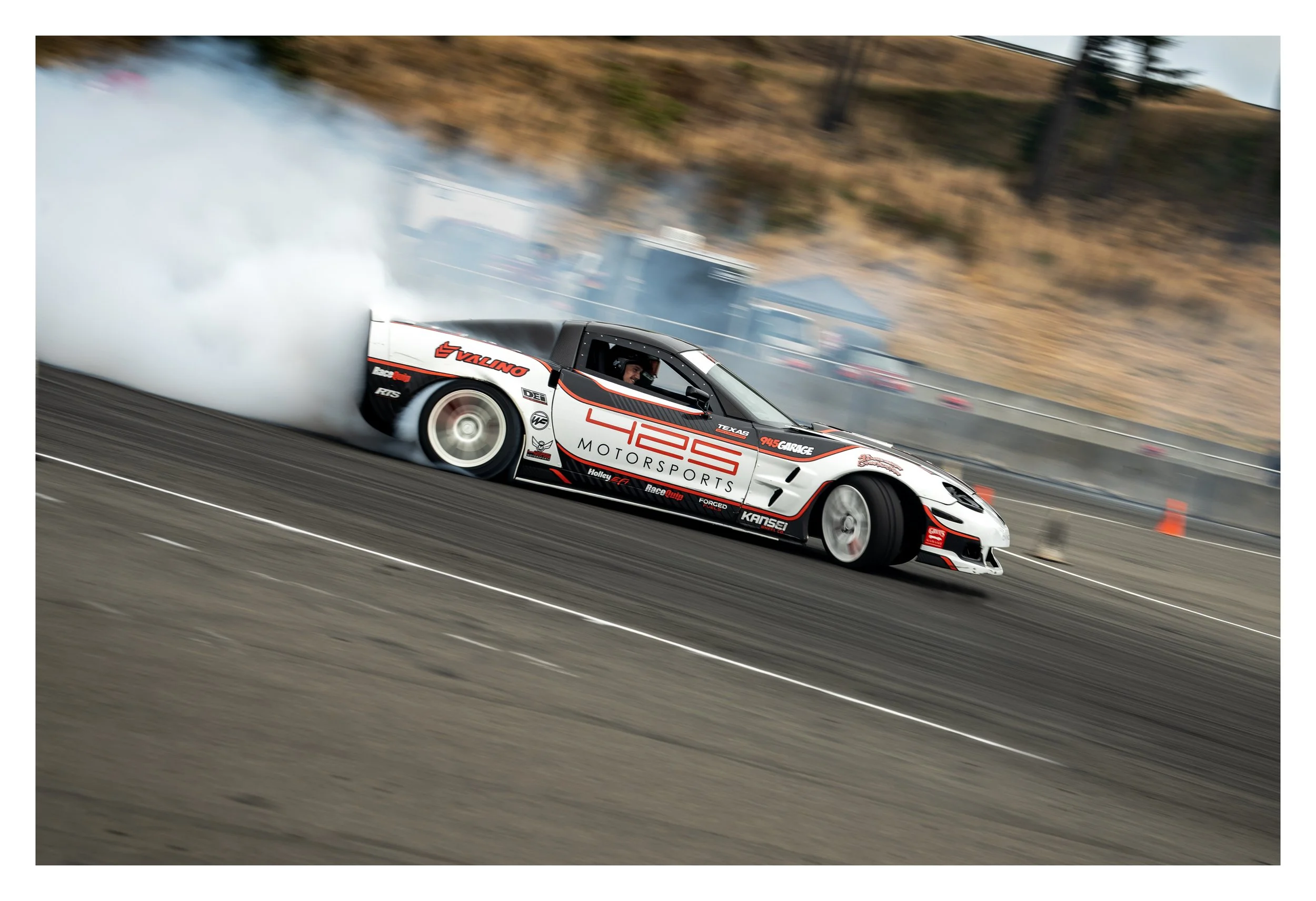 A race car performing a burnout on a racetrack with tire smoke, hills, and trees in the background.