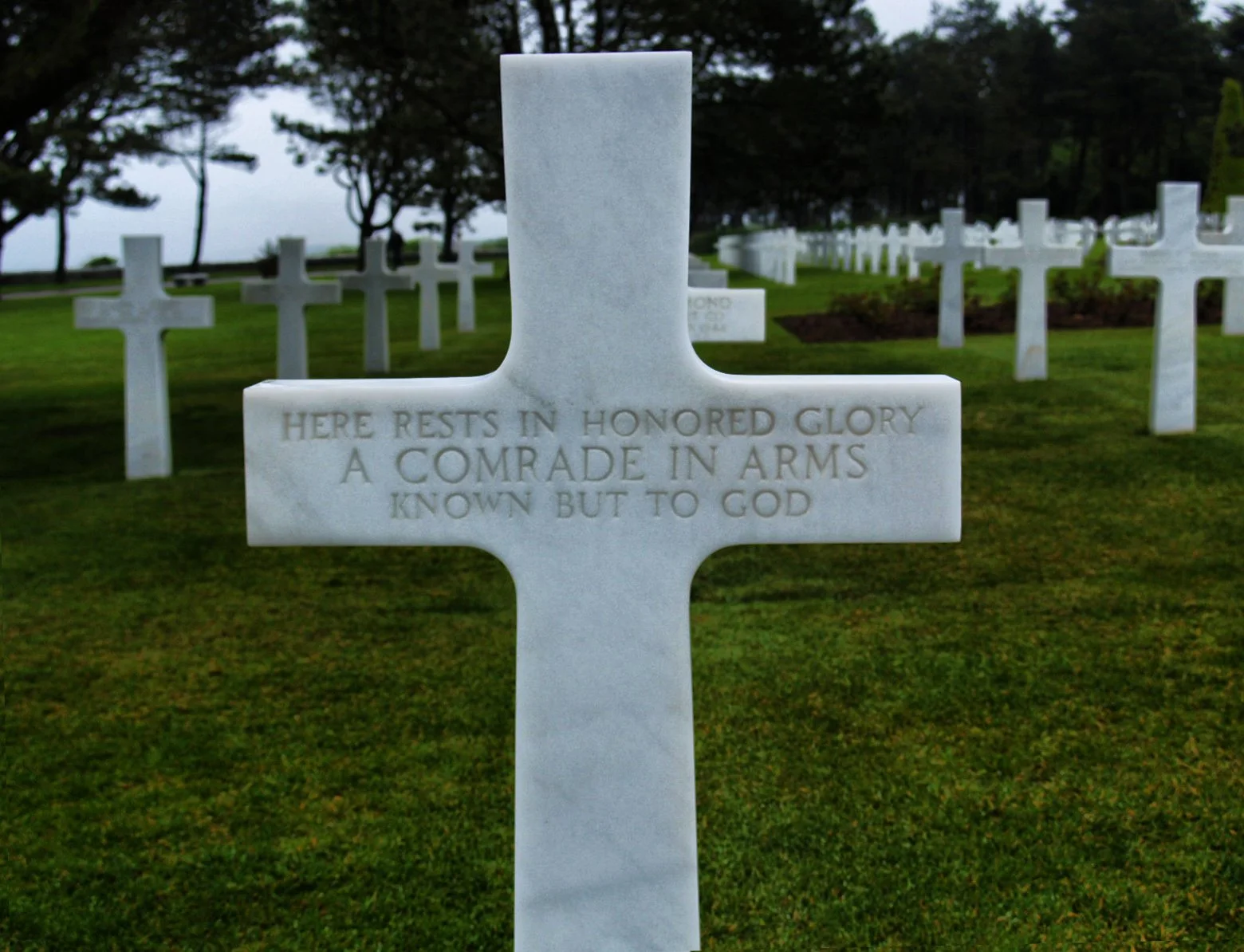 White marble cross headstone in a military cemetery with the inscription: 'Here rests in honored glory a comrade in arms known but to God,' surrounded by other similar headstones and trees in the background.