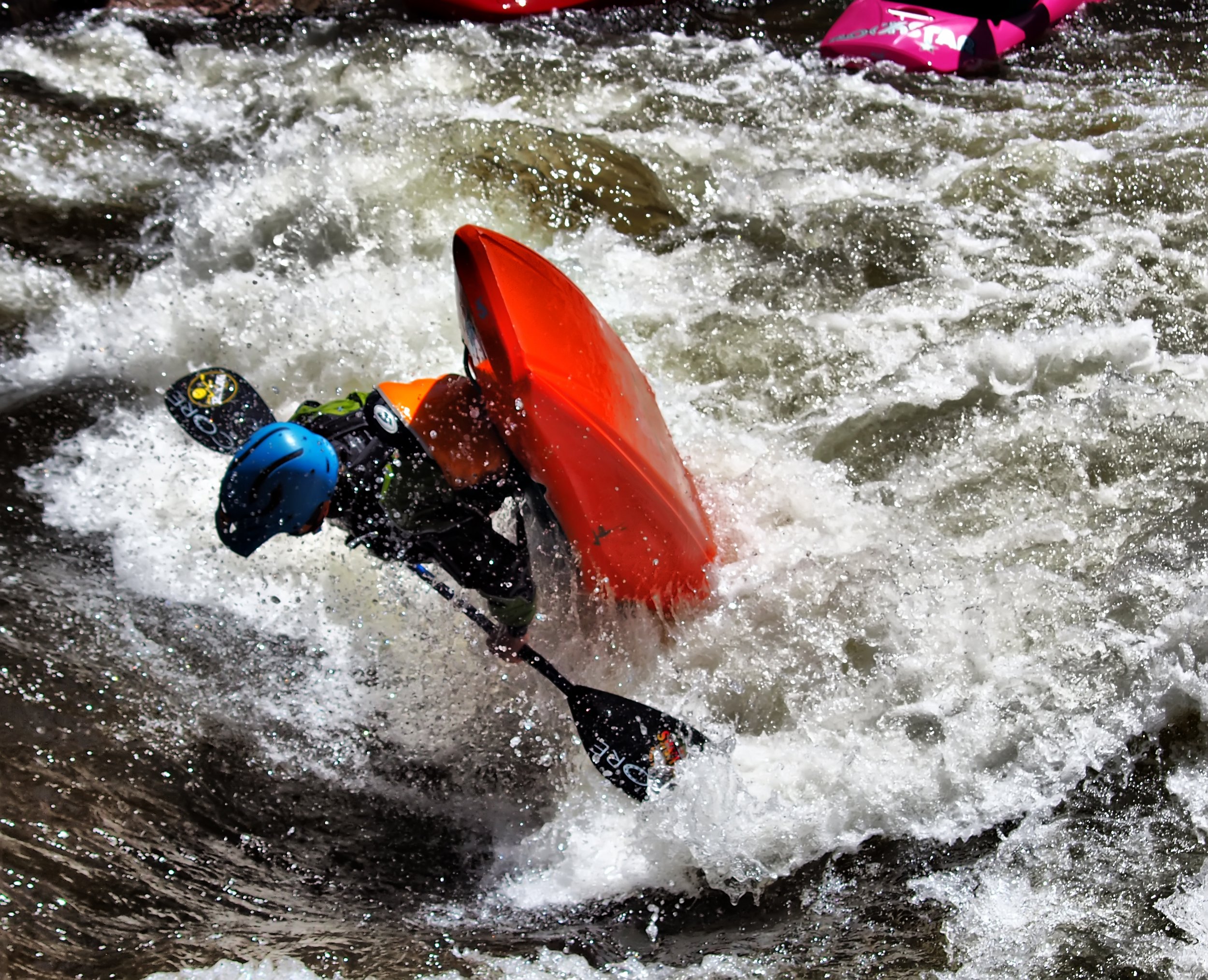 A person kayaking in fast-moving, white-water rapids with a red kayak and a blue helmet.