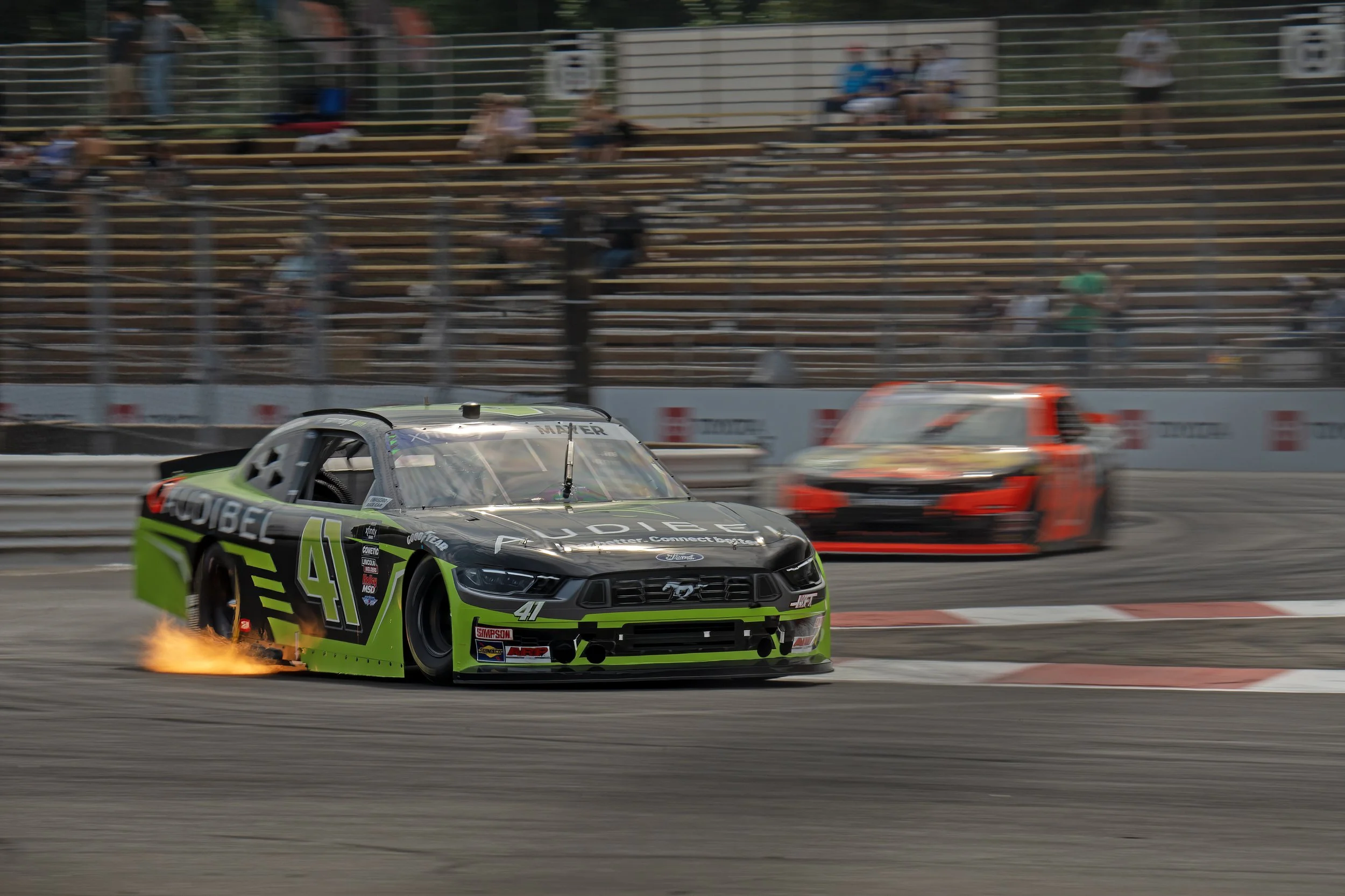 NASCAR race car crashing as flames and sparks fly from the rear at a racetrack with empty grandstands in the background.