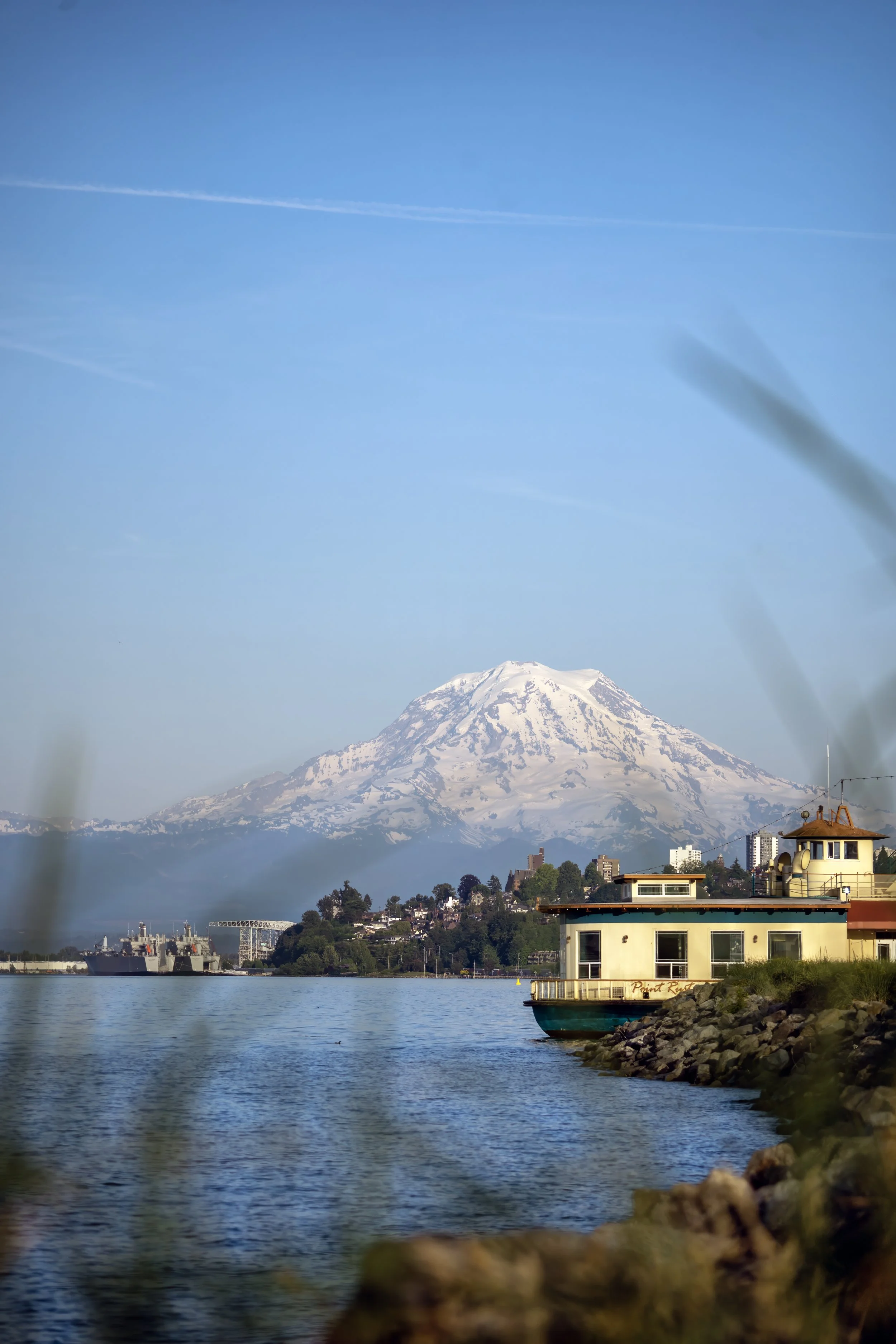 View of Mount Rainier with snow, seen across a body of water. Buildings and a ship are in the foreground, and the sky is mostly clear with a few wispy clouds.