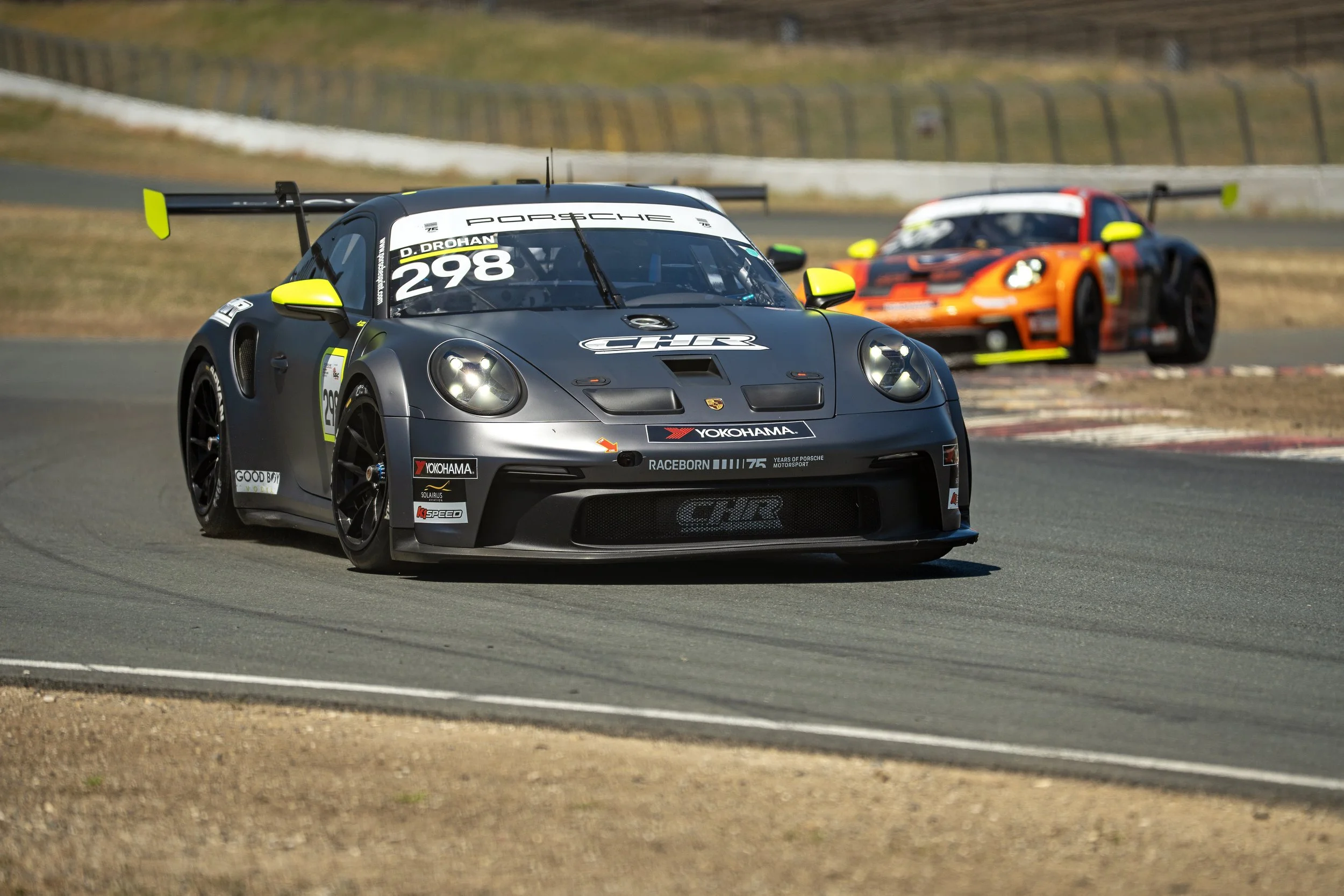 A black race car with yellow accents driving on a racetrack, with an orange and black race car in the background.
