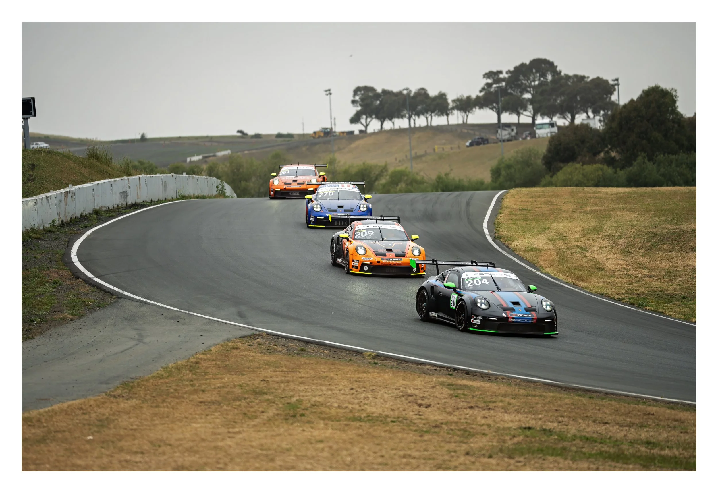 Race cars on a curving race track with trees and parked vehicles in the background.