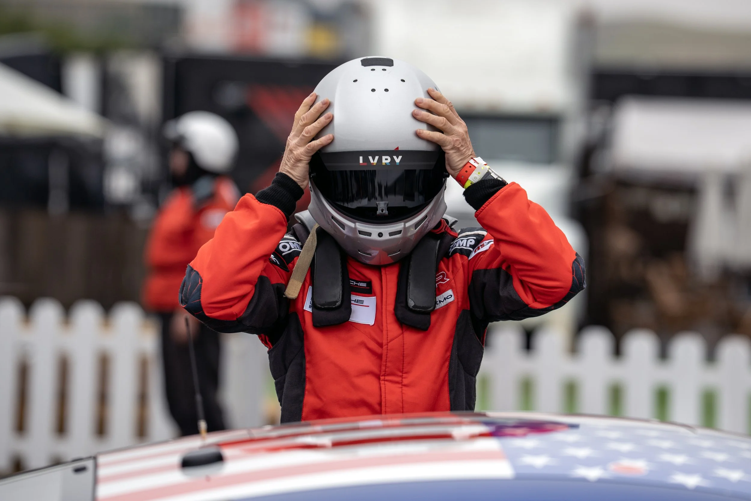 Person wearing racing gear and helmet adjusting helmet, with race car in front.