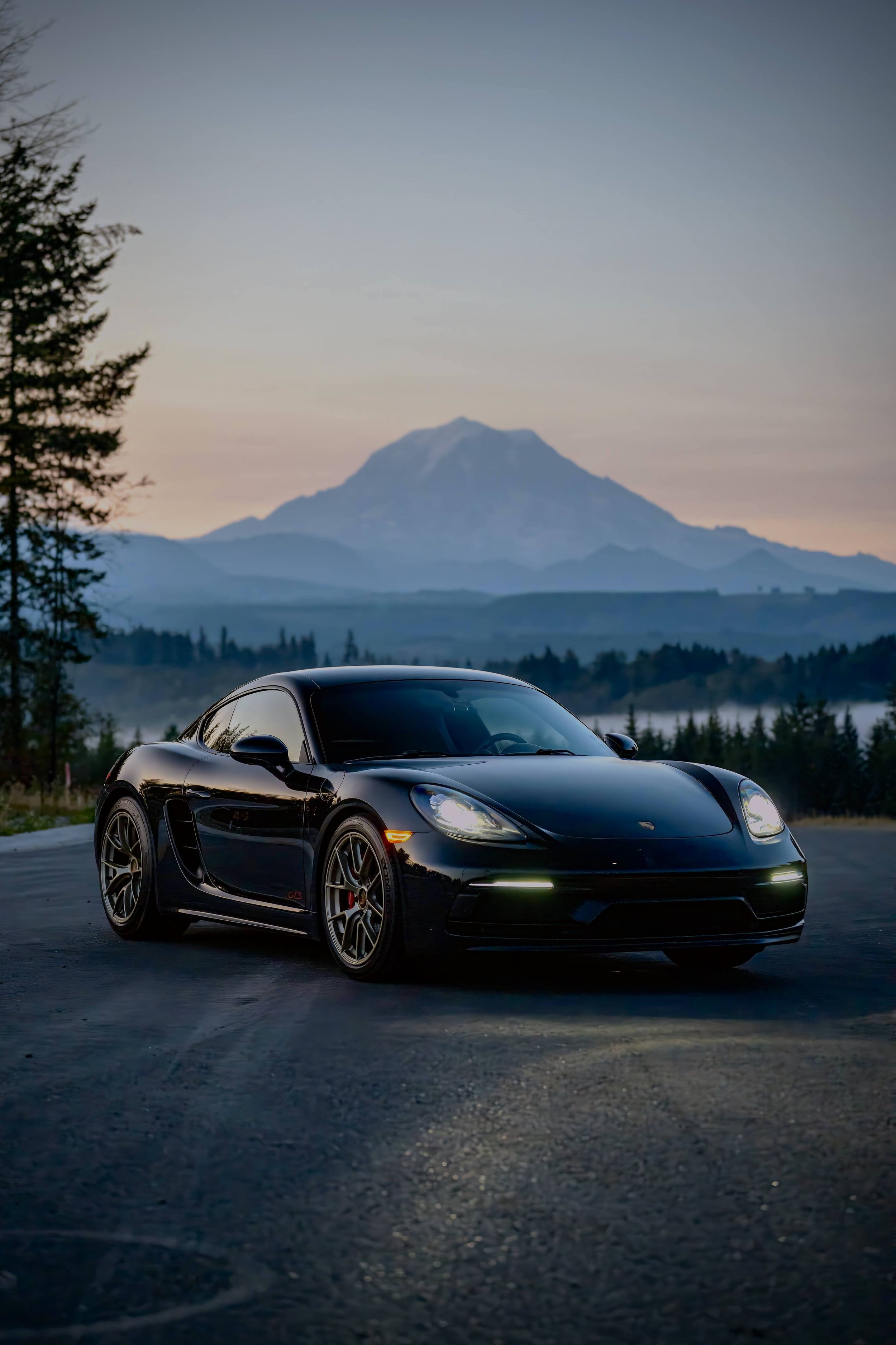 A black sports car parked on an empty road during sunset with mountains and a forest in the background.