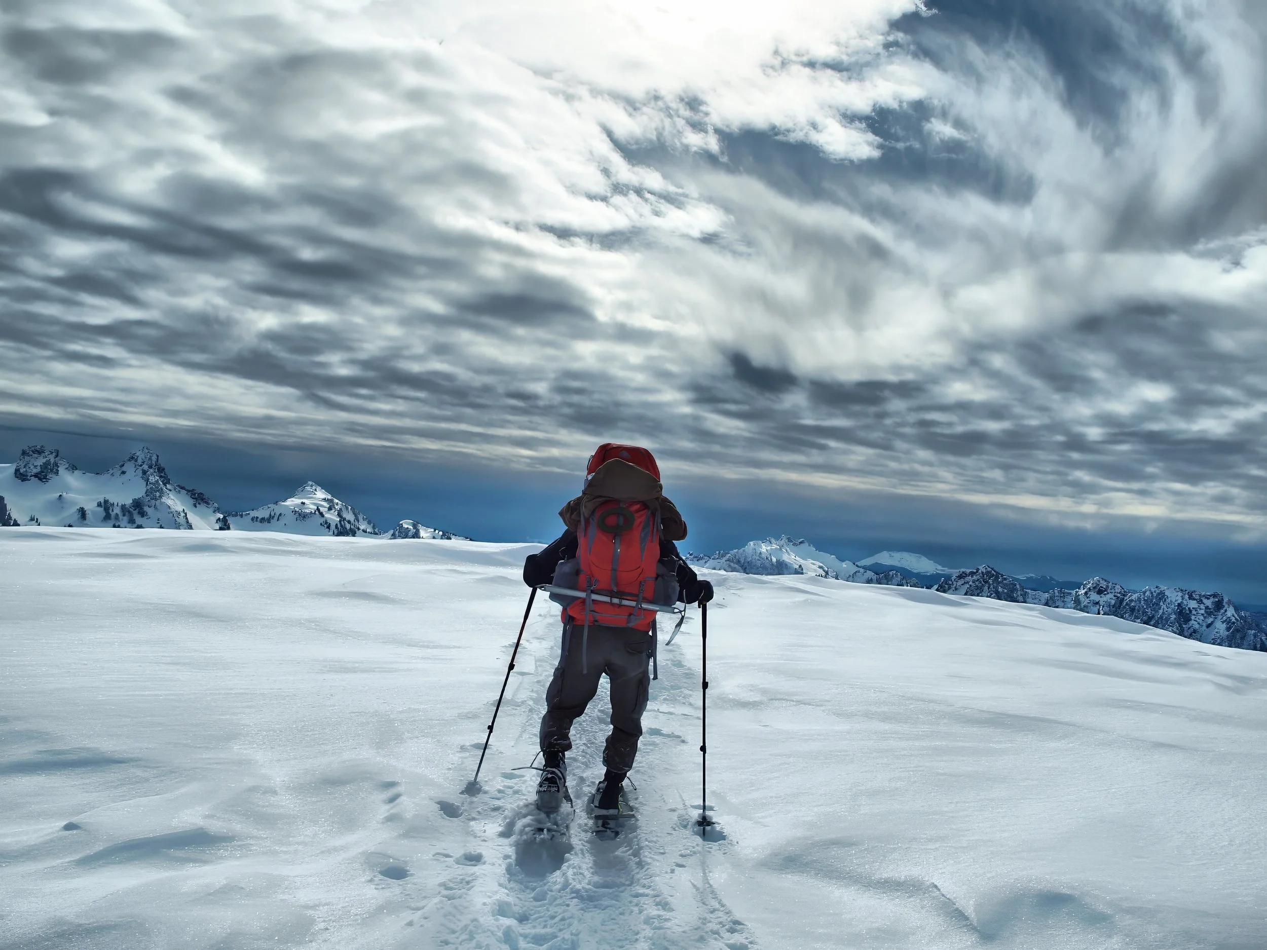 A person dressed in winter gear, including a red backpack and trekking poles, snowshoeing across a snowy, mountainous landscape under a cloudy sky.
