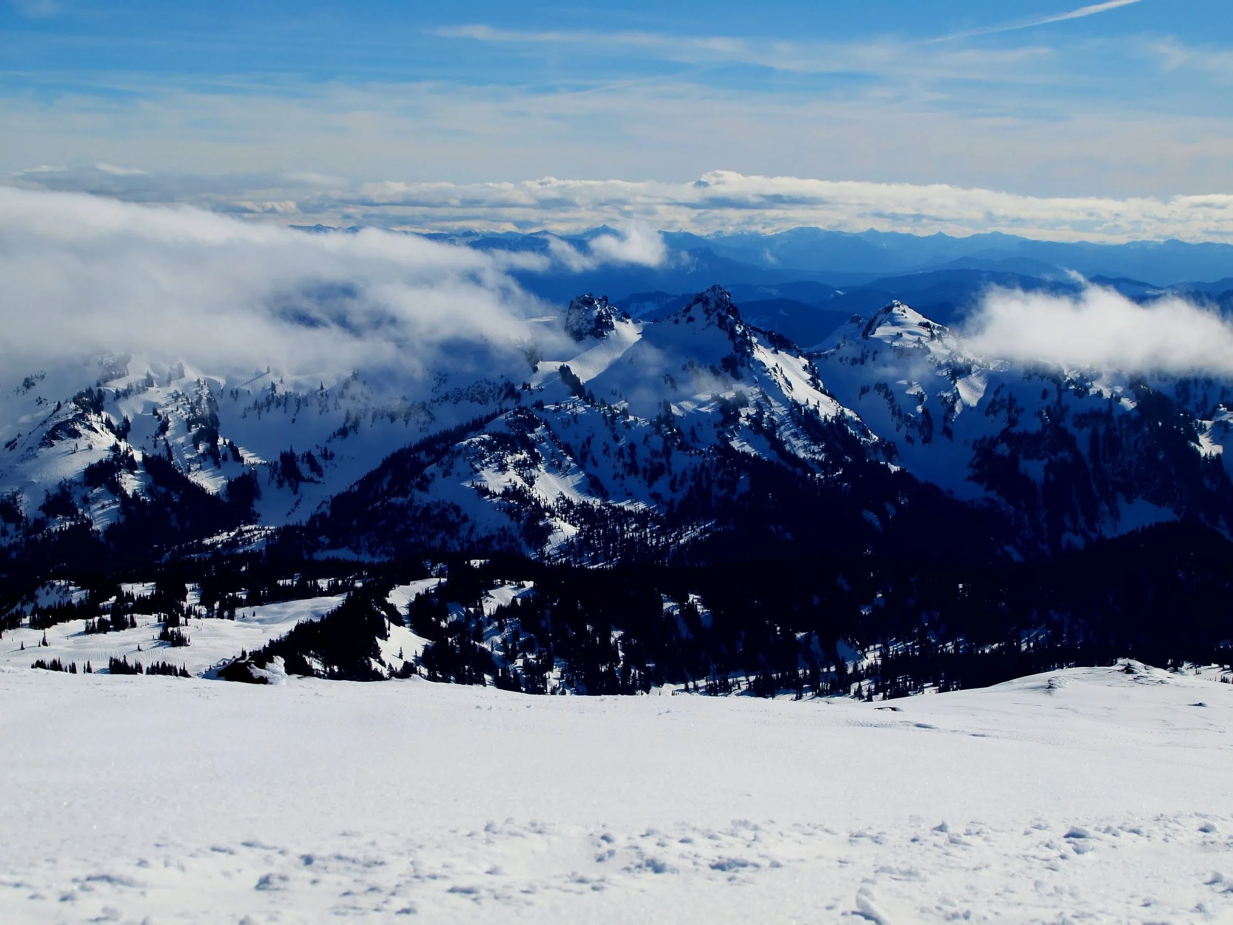 Snow-covered mountain landscape with rugged peaks, covered with snow, surrounded by clouds and a partly cloudy blue sky.