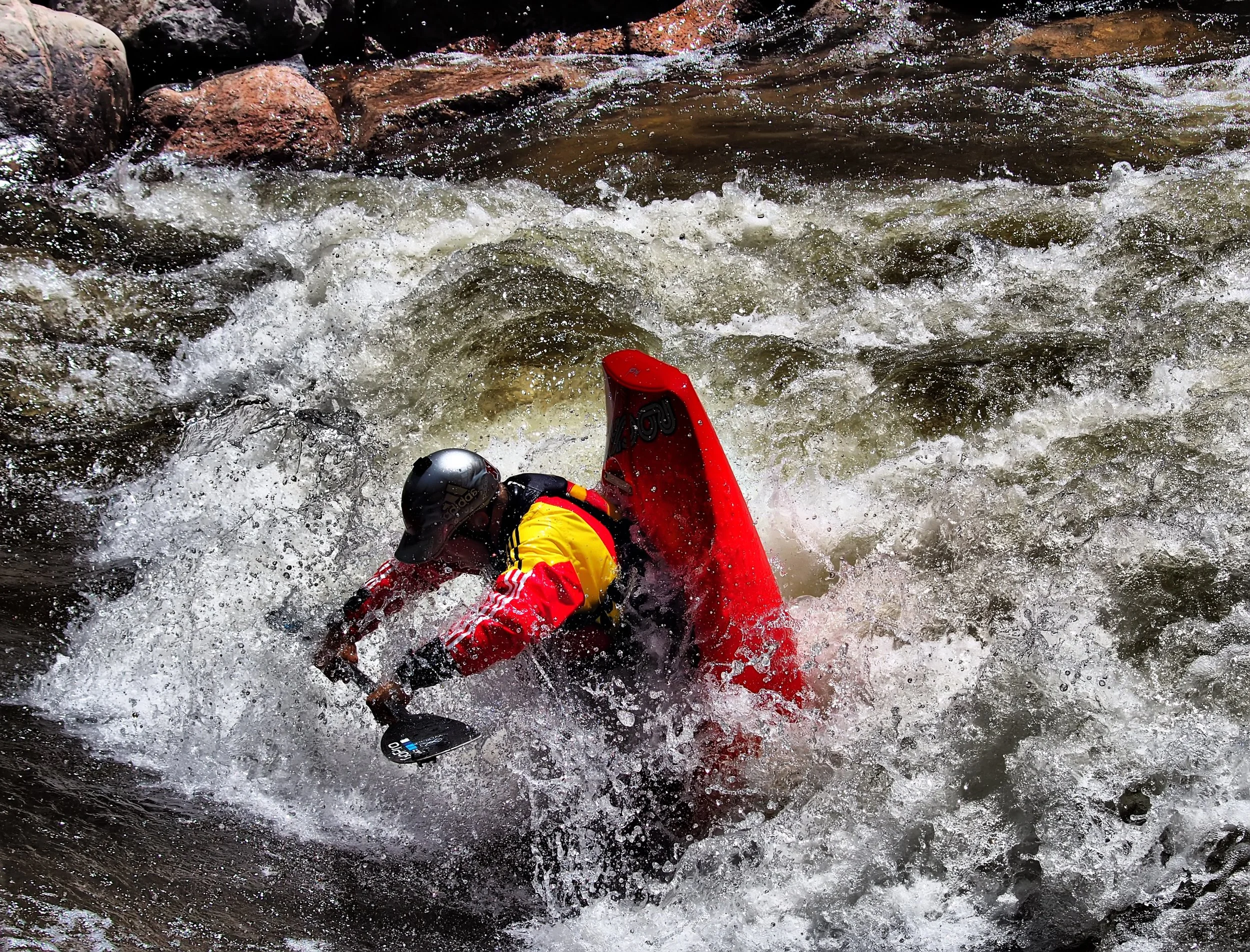 A person in a red kayak wearing a helmet is navigating through white water rapids.