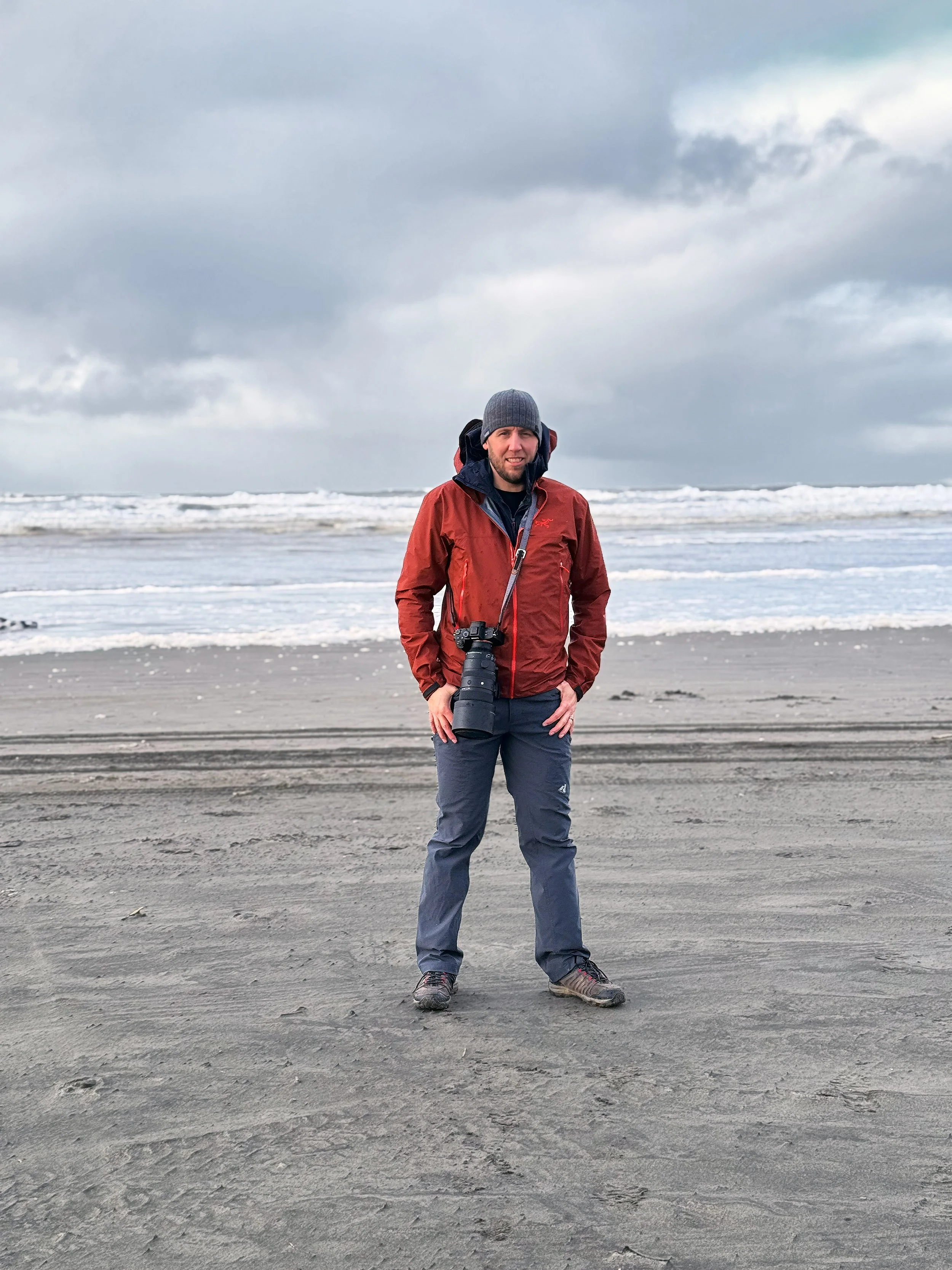 Matthew Zalewski wearing a red jacket and carrying his camera on a beach in the Pacific Northwest