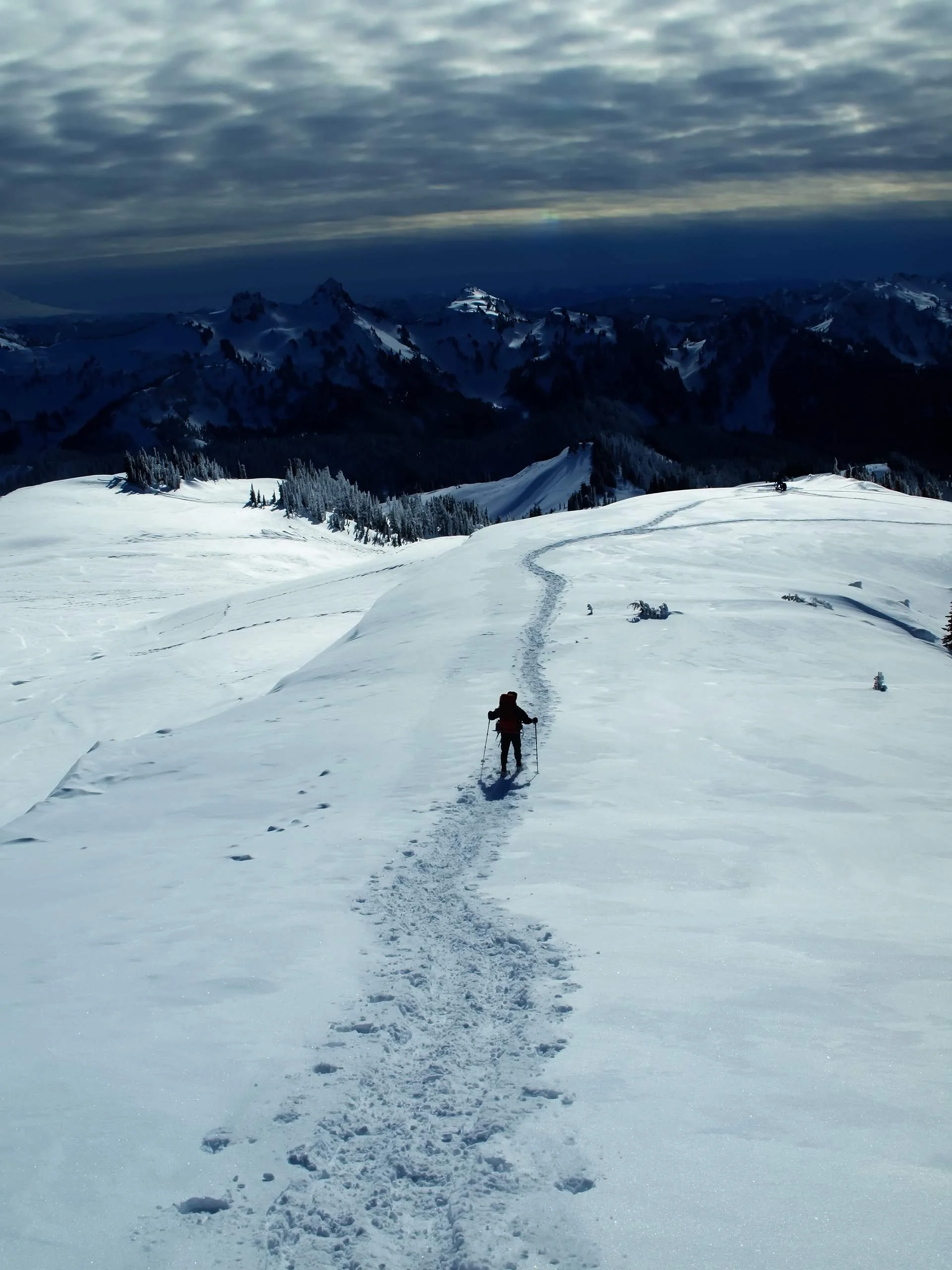 Person walking through snowy mountain landscape with ski poles, with tracks in the snow leading toward distant mountain peaks under cloudy sky.