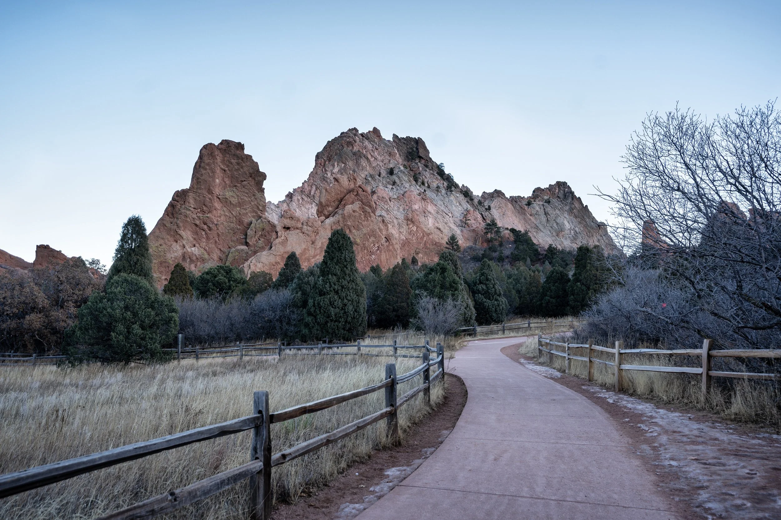 A scenic view of rocky mountains in the background with a winding paved trail, green trees, and a wooden fence in the foreground.