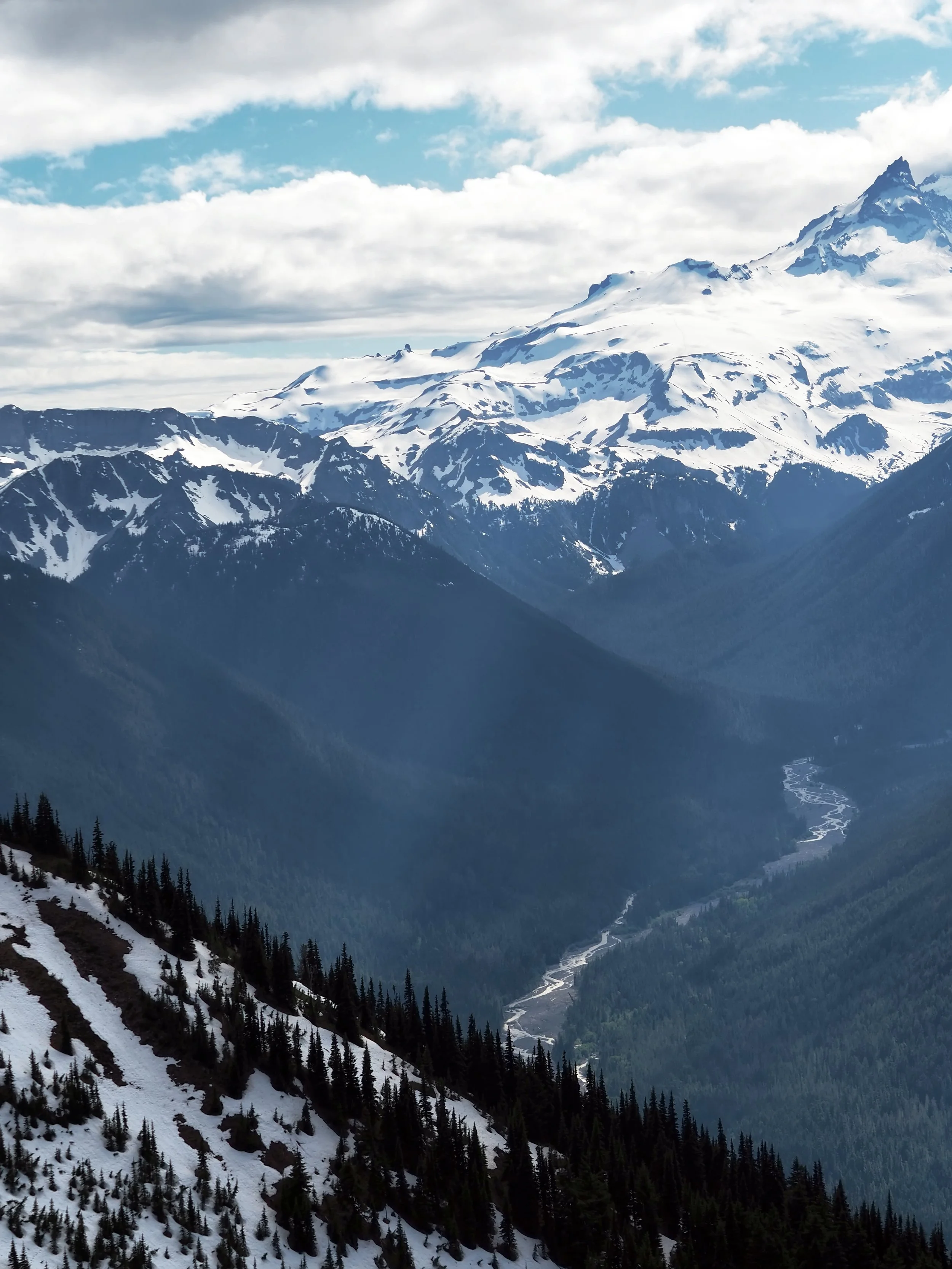 Snow-covered mountain range with pine trees on the slopes and a partly cloudy sky.