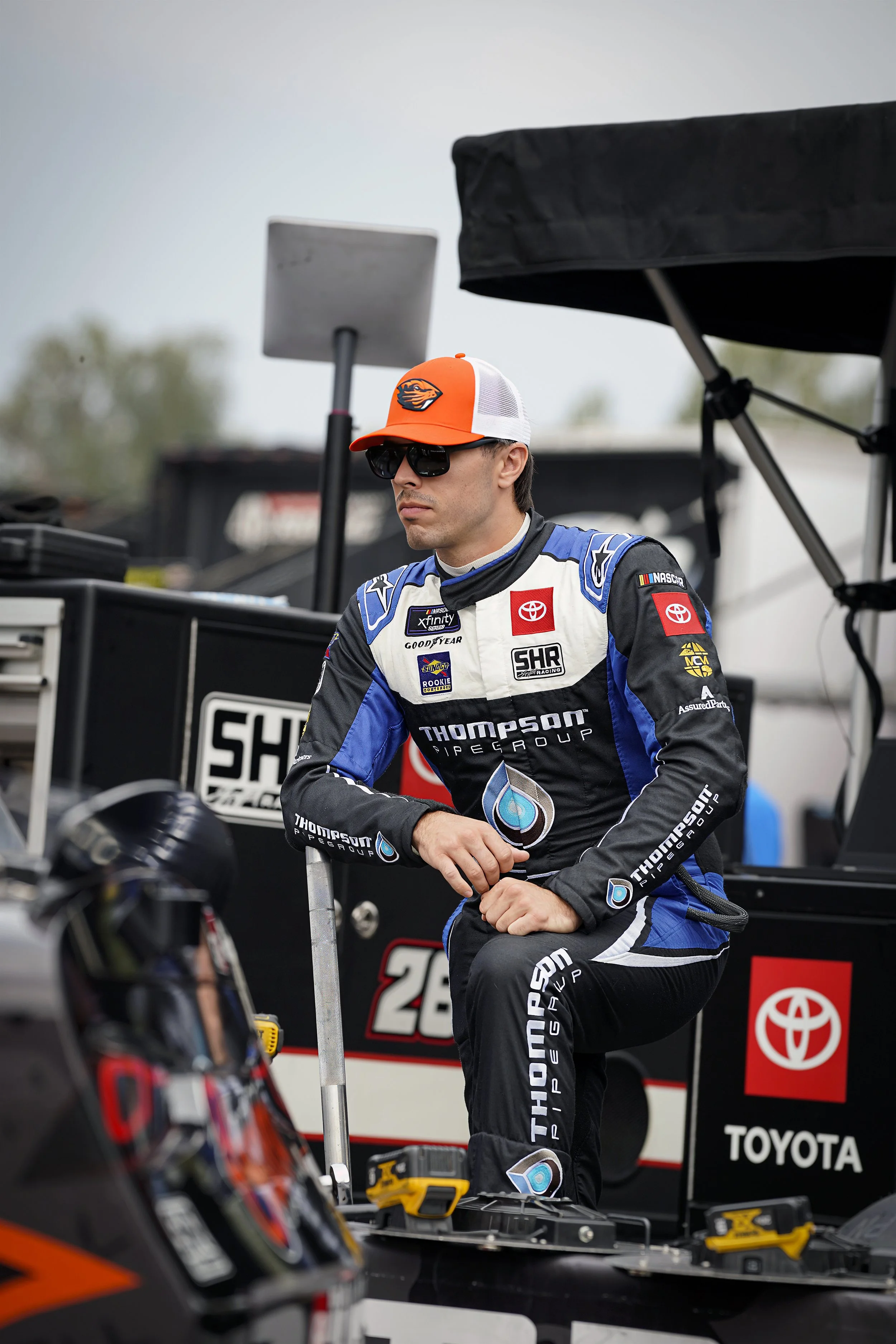 Race car driver in racing suit standing near pit equipment with race car in foreground, team trailer and tools in background.