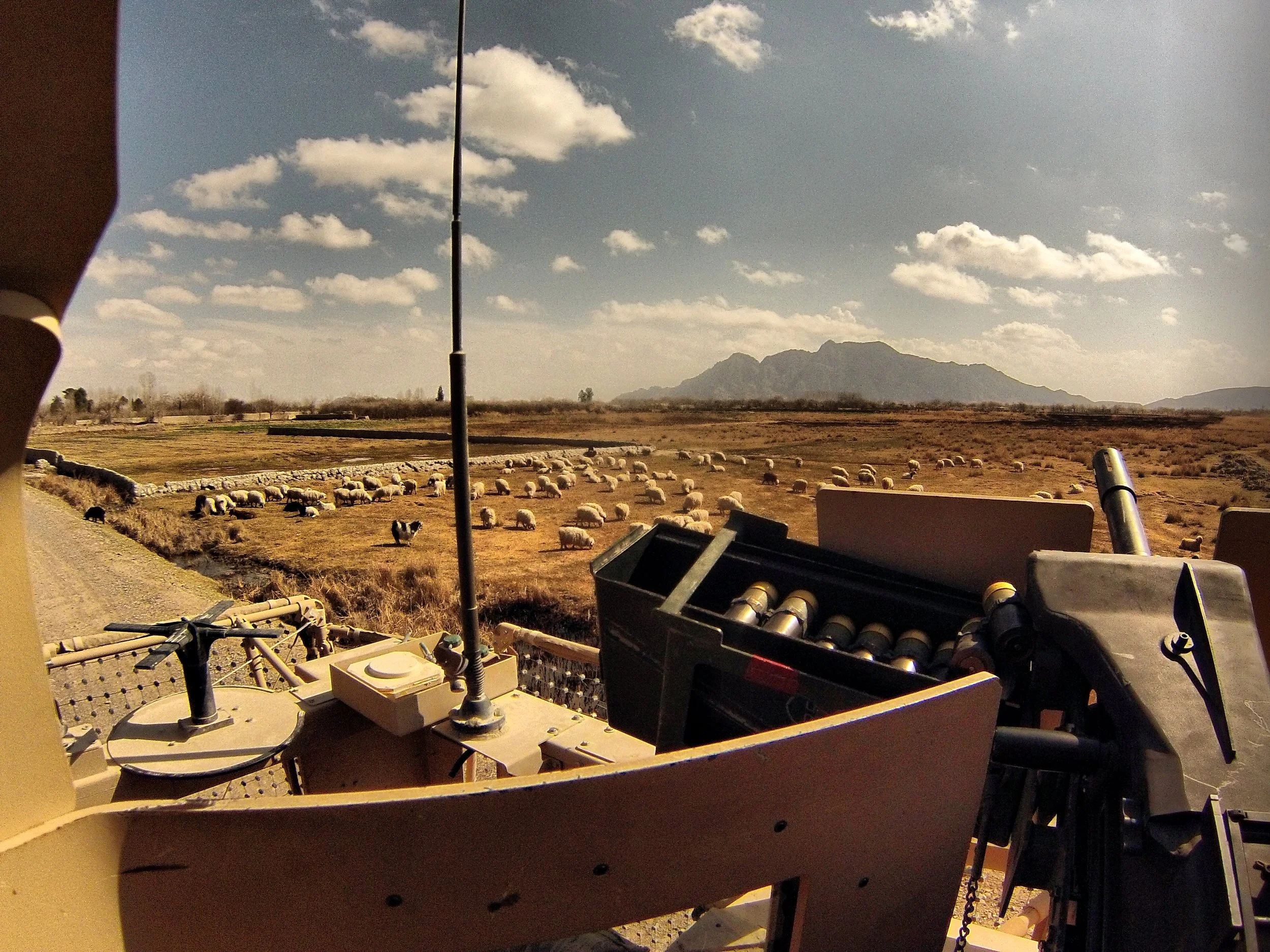 View from a military vehicle's turret overlooking a rural landscape with sheep grazing and mountains in the distance.