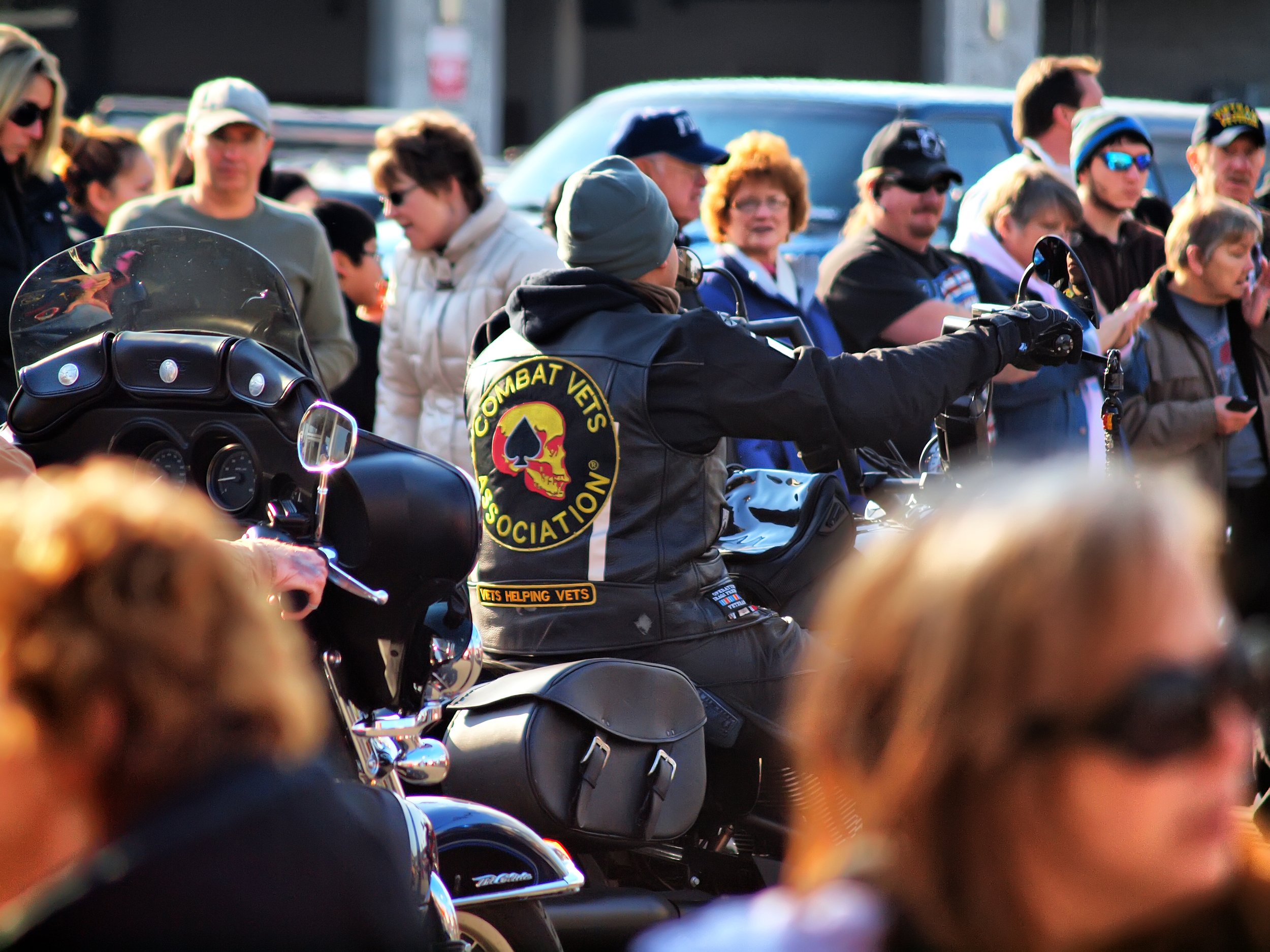 A group of people gathered around a motorcyclist wearing a black leather vest with a 'Combat Veterans Motorcycle Association' patch and a skull logo. The scene appears to be outdoors during daylight.