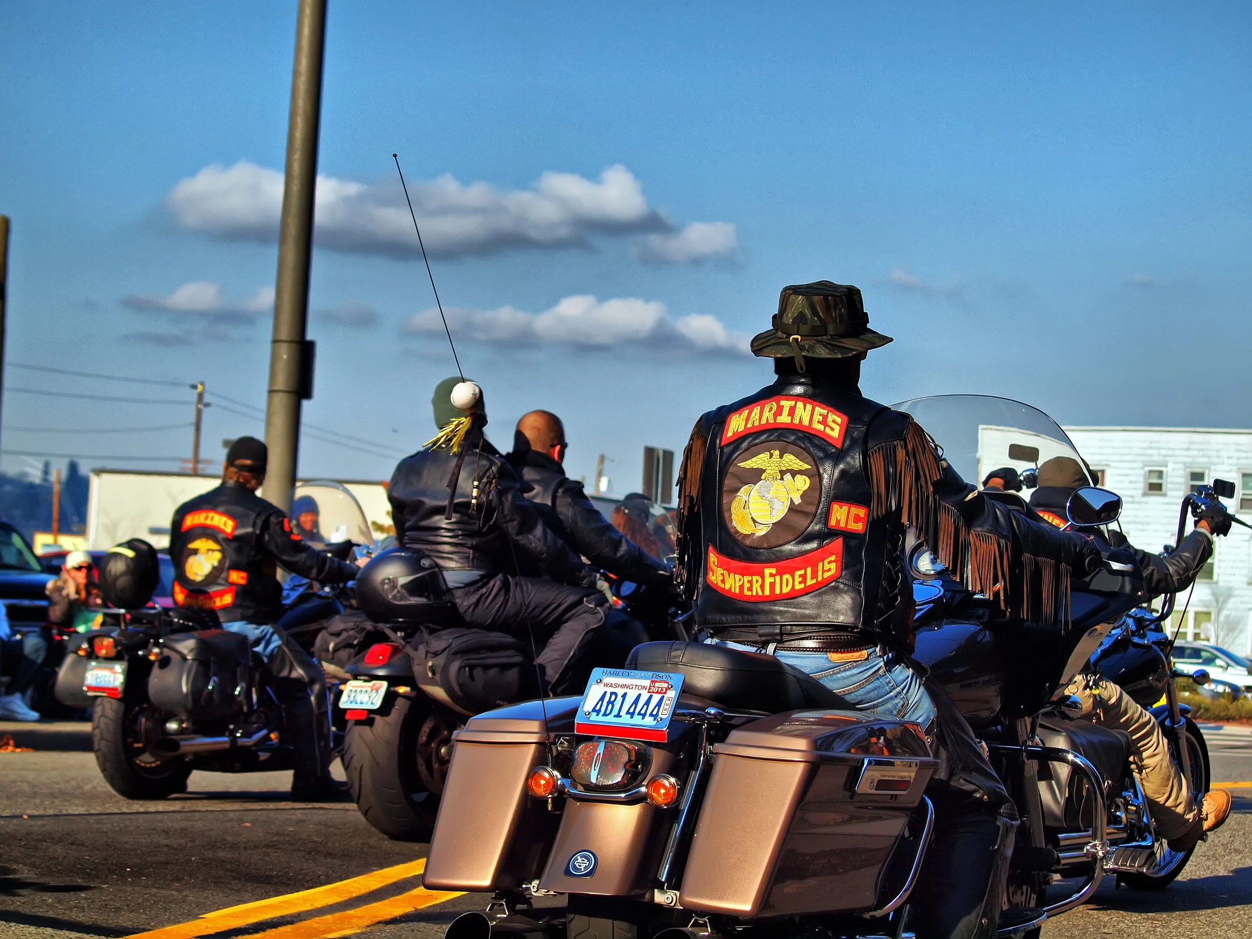 A group of motorcycle riders, including members of the Marines, are gathering on a street during daytime, with some wearing Marine jackets and insignia, and others in black leather jackets and helmets.