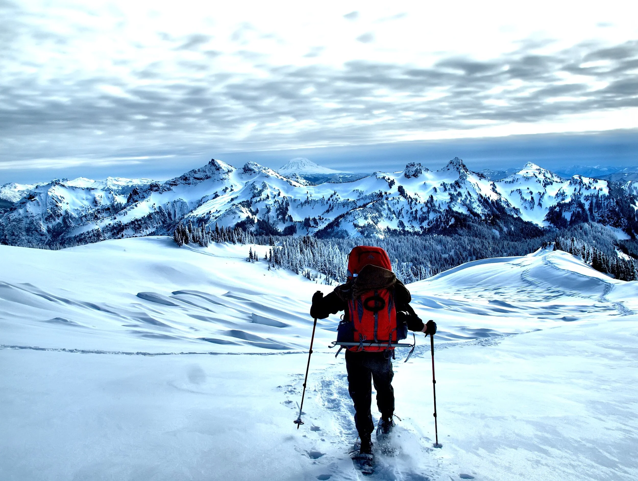A person dressed in winter gear is sitting on a sled in a snowy mountainous landscape, with snow-covered peaks and cloudy sky in the background.